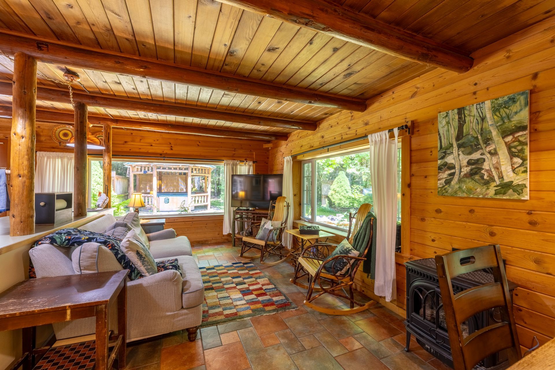 Cozy living room in a log cabin with wood walls, ceiling, and floors. Furnishings include sofas, rocking chairs, and a small wood-burning stove, with large windows overlooking greenery.