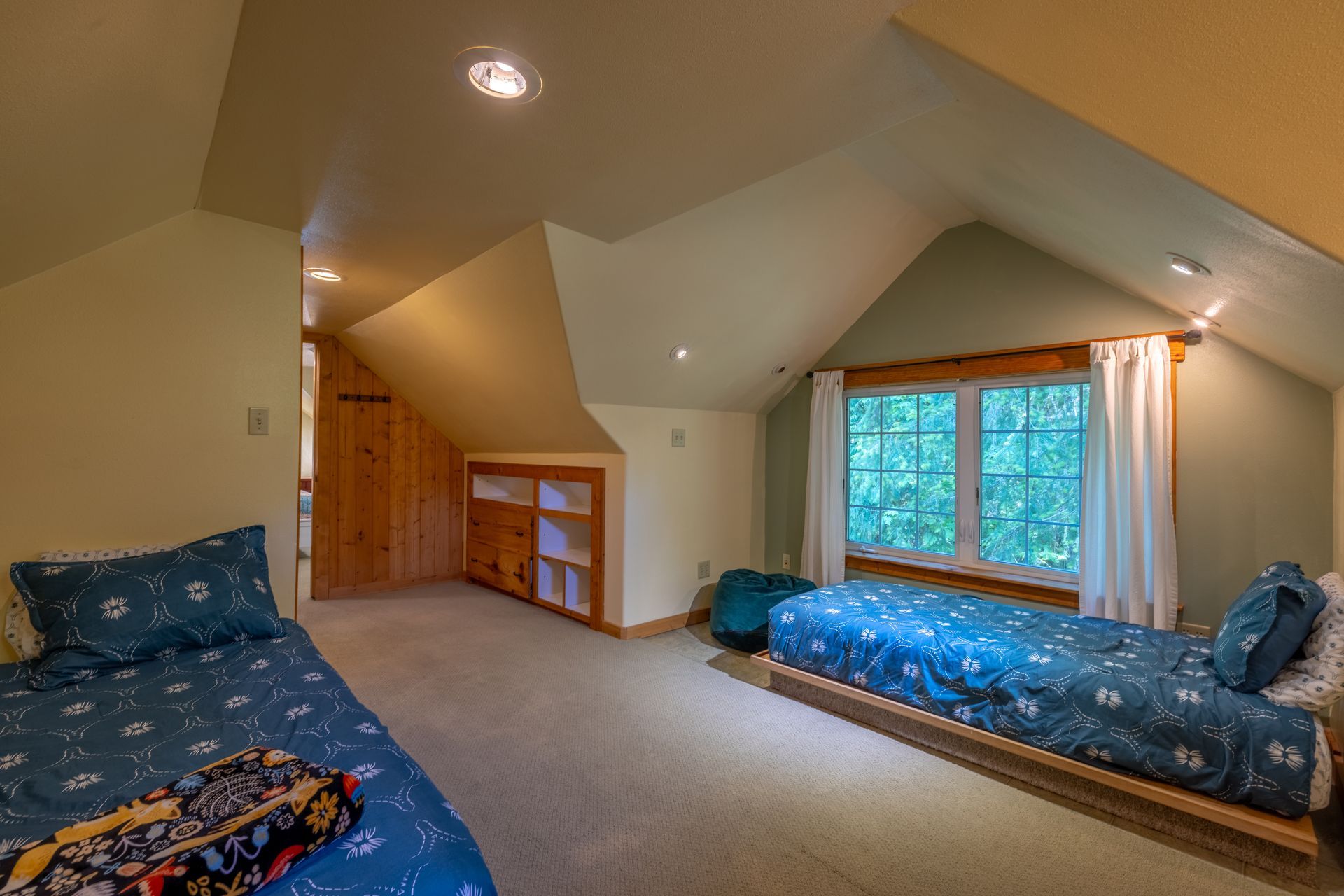 Bedroom with two twin beds, a built-in bookcase, and a window looking out to trees. Walls are light-colored and the beds have blue patterned bedding.