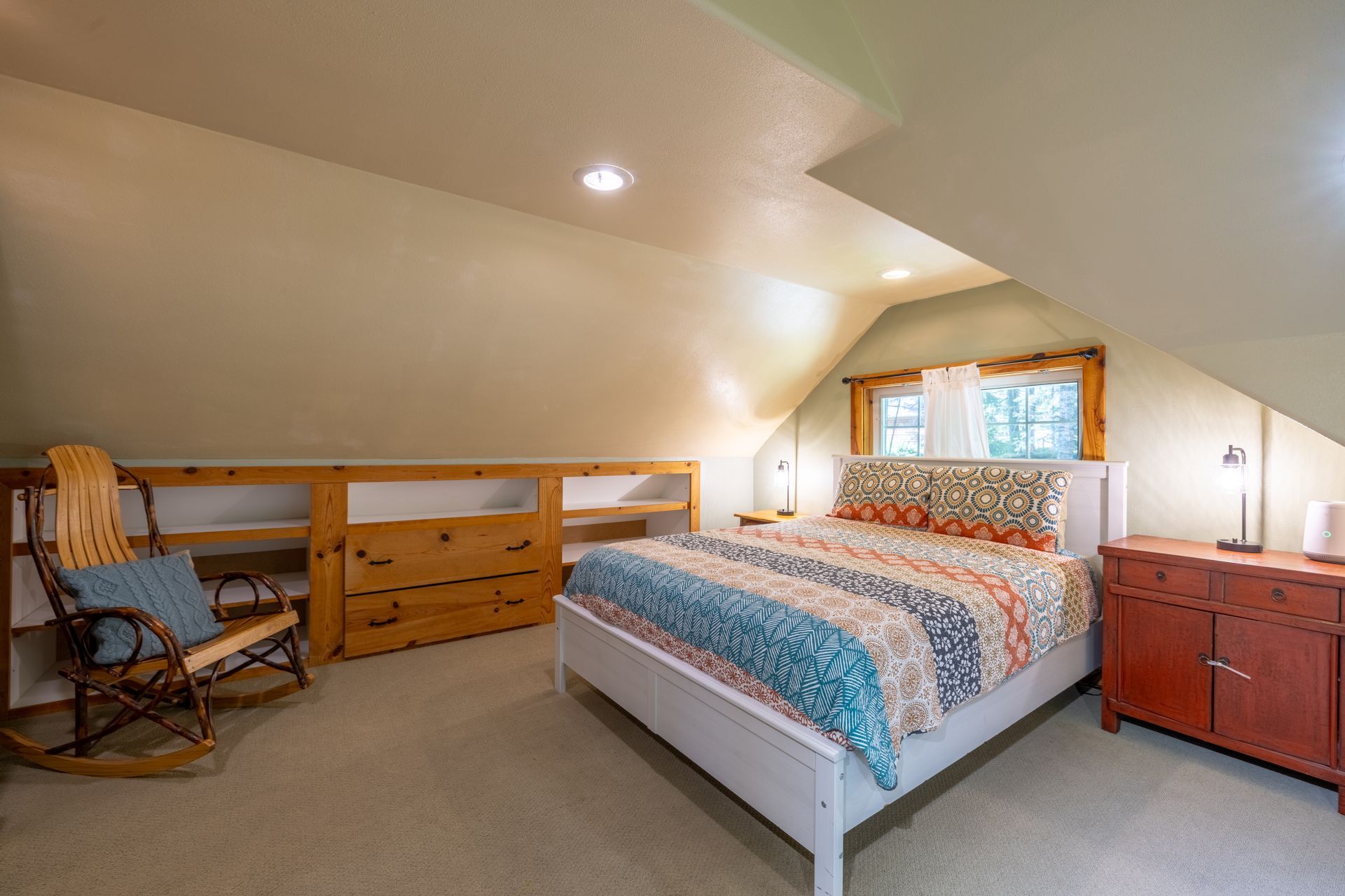 Cozy attic bedroom with a bed, dresser, rocking chair, and built-in shelves, bathed in natural light.