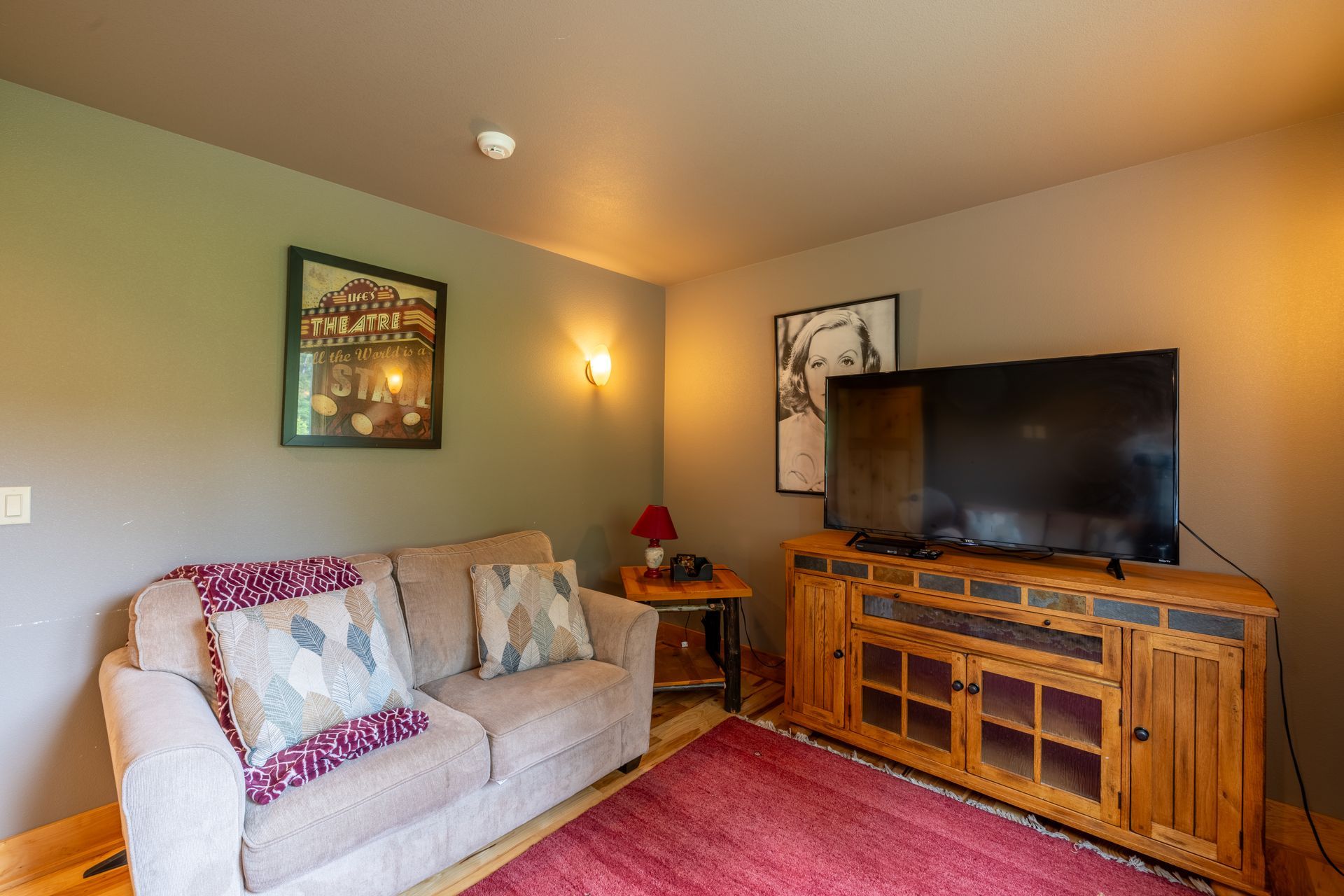 Living room with a beige couch, wooden entertainment center with a TV, and red rug. The walls are gray.