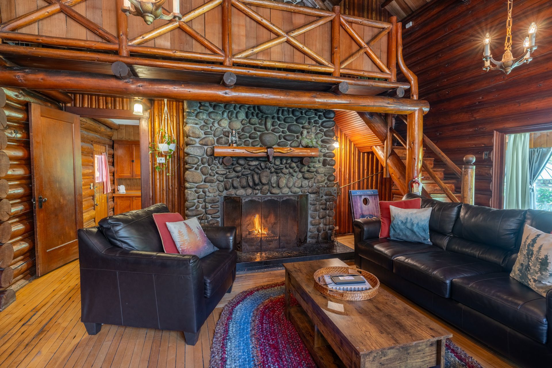 Cozy cabin living room with a stone fireplace, leather furniture, wooden beams, and a loft.