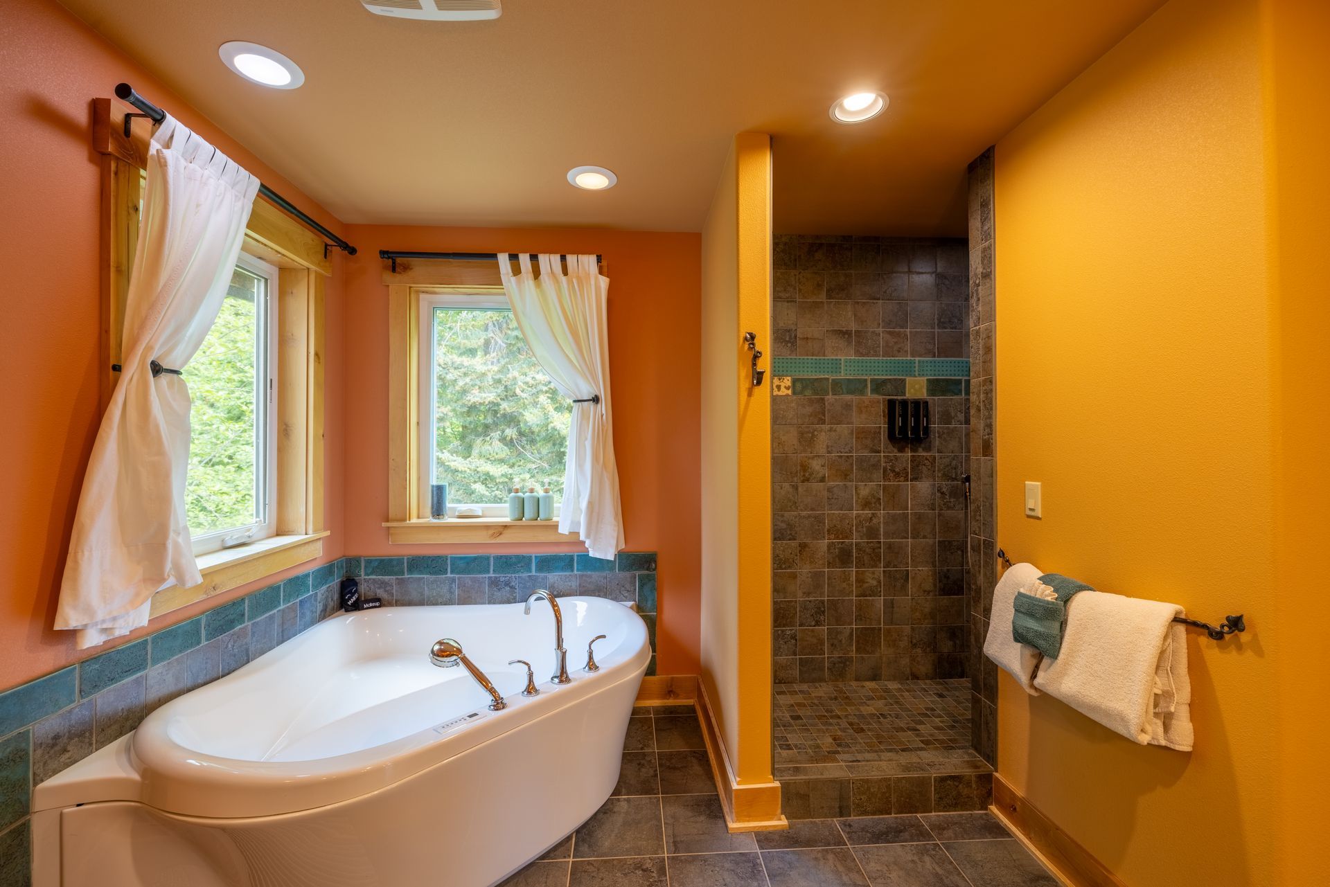 Bathroom with a freestanding white tub next to two windows with sheer curtains, a walk-in shower, and colorful orange and teal accents.