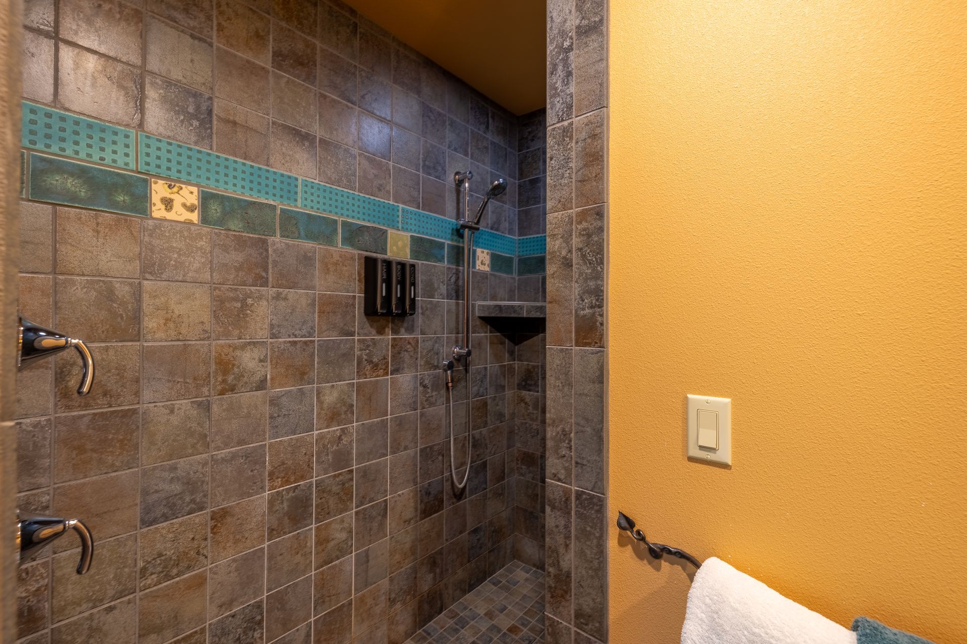Tiled shower with brown and teal accents, a handheld shower head, and a towel hanging on the adjacent wall.