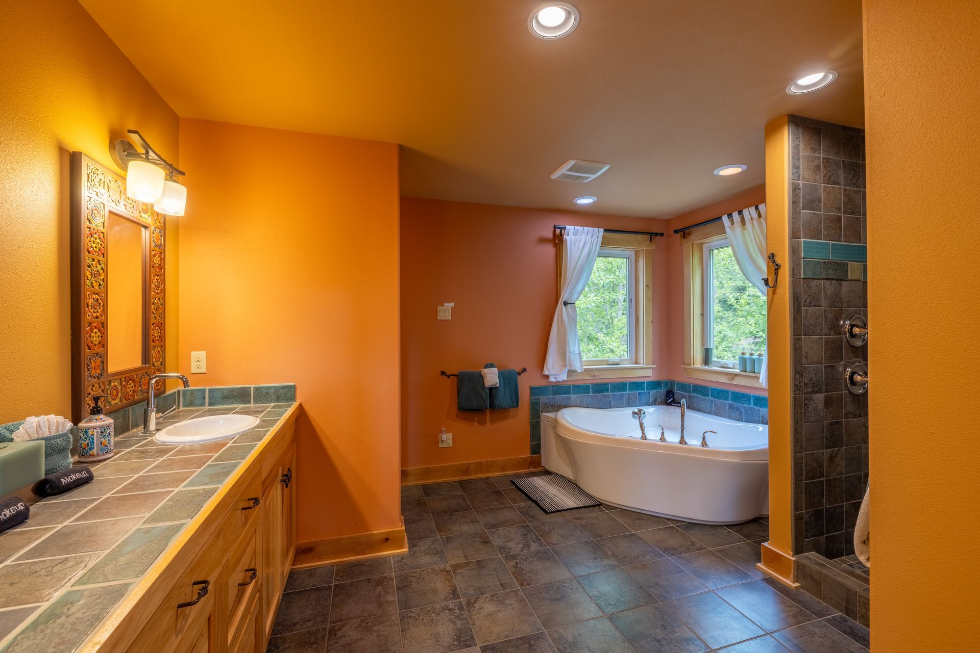 Bathroom with orange walls, wooden vanity with two sinks, a corner bathtub with blue tile surround, and a dark tiled shower.