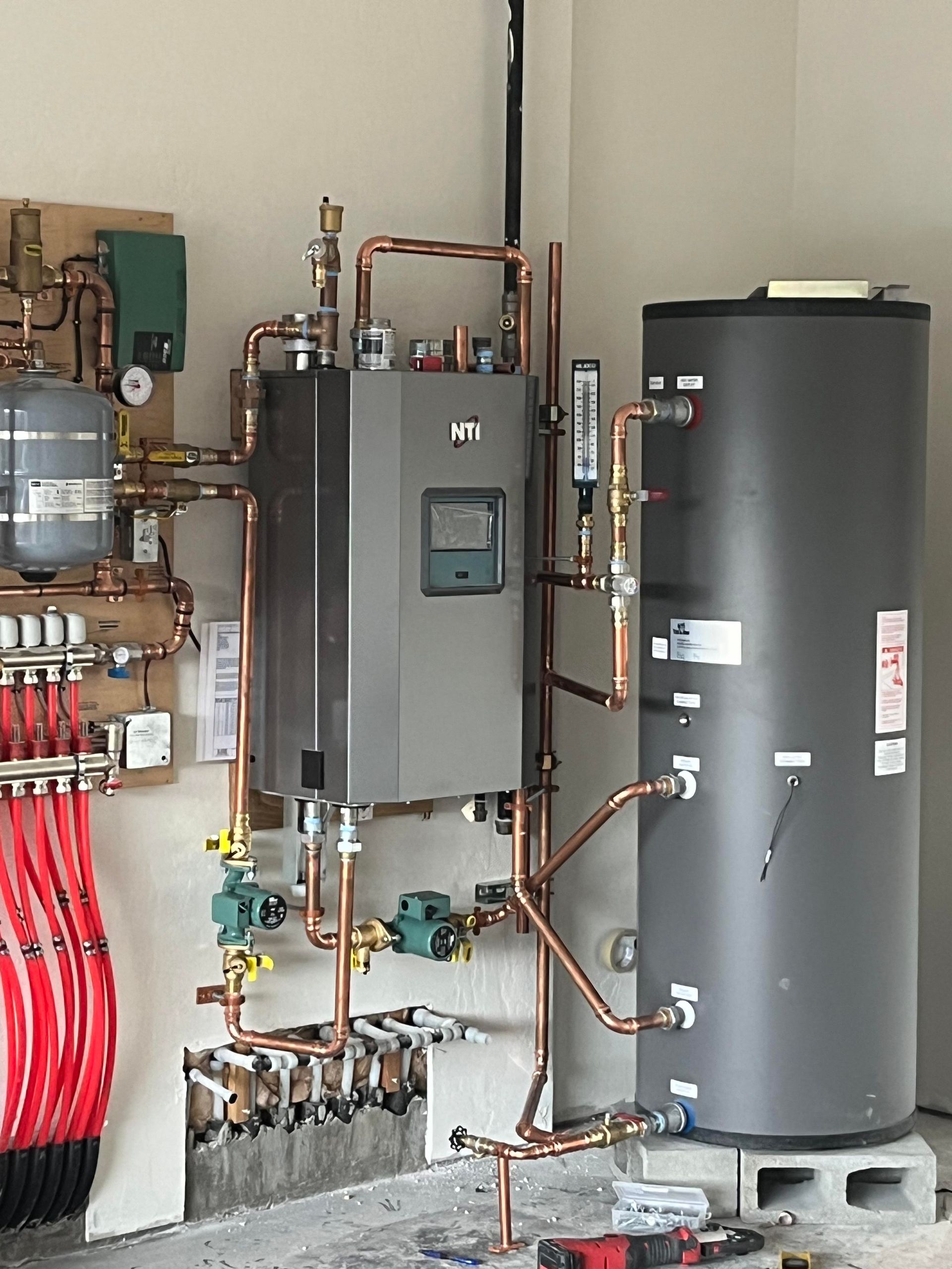 Heating system with a grey boiler and water tank, connected by copper pipes, mounted on a concrete block.