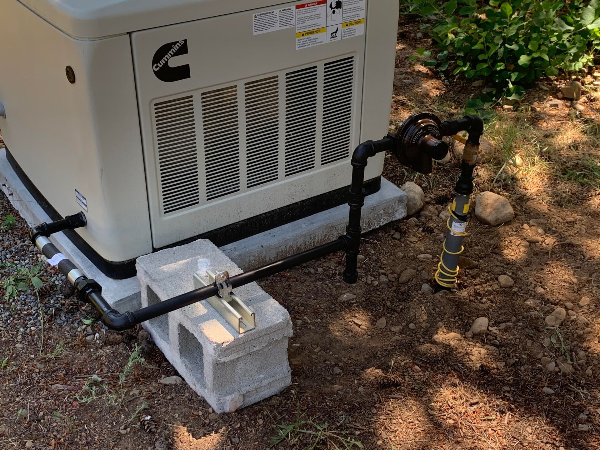 A tan Cummins generator on cinder blocks with black gas piping, outdoors.