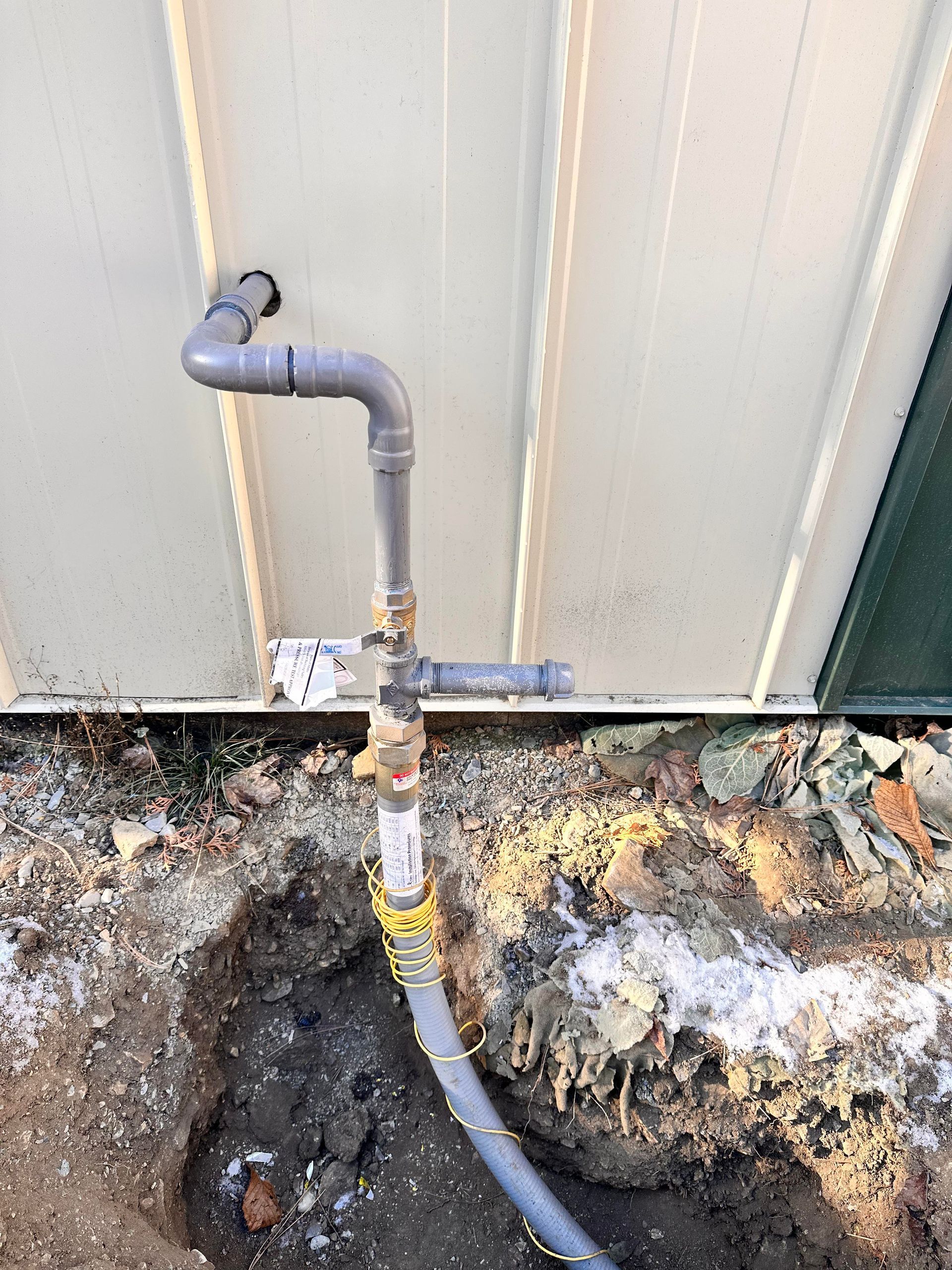 Outdoor plumbing, grey pipe with fittings, connected to a building with dirt around the base.