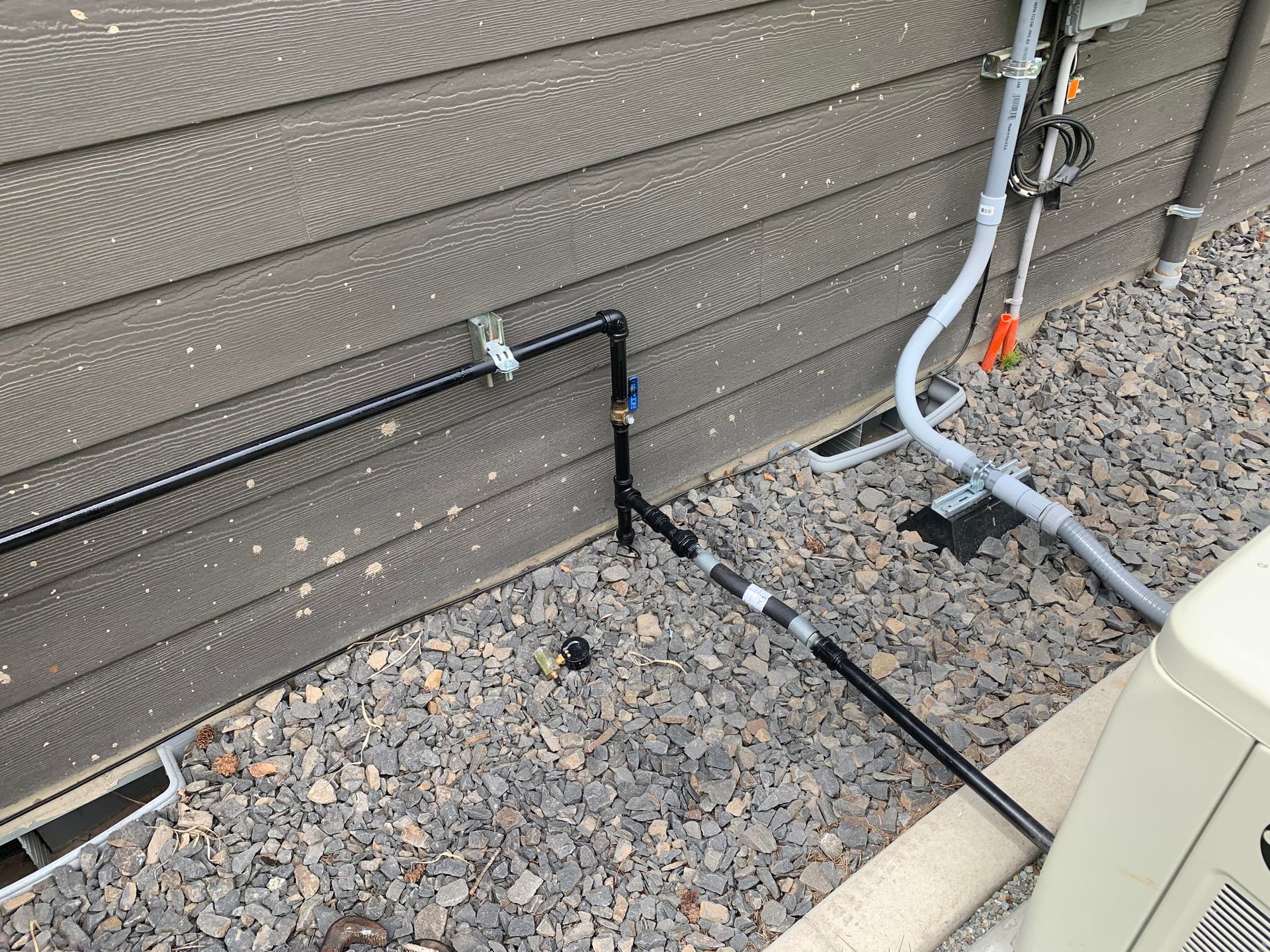 Black gas pipes along a brown house exterior, connecting to an appliance in a gravel yard.