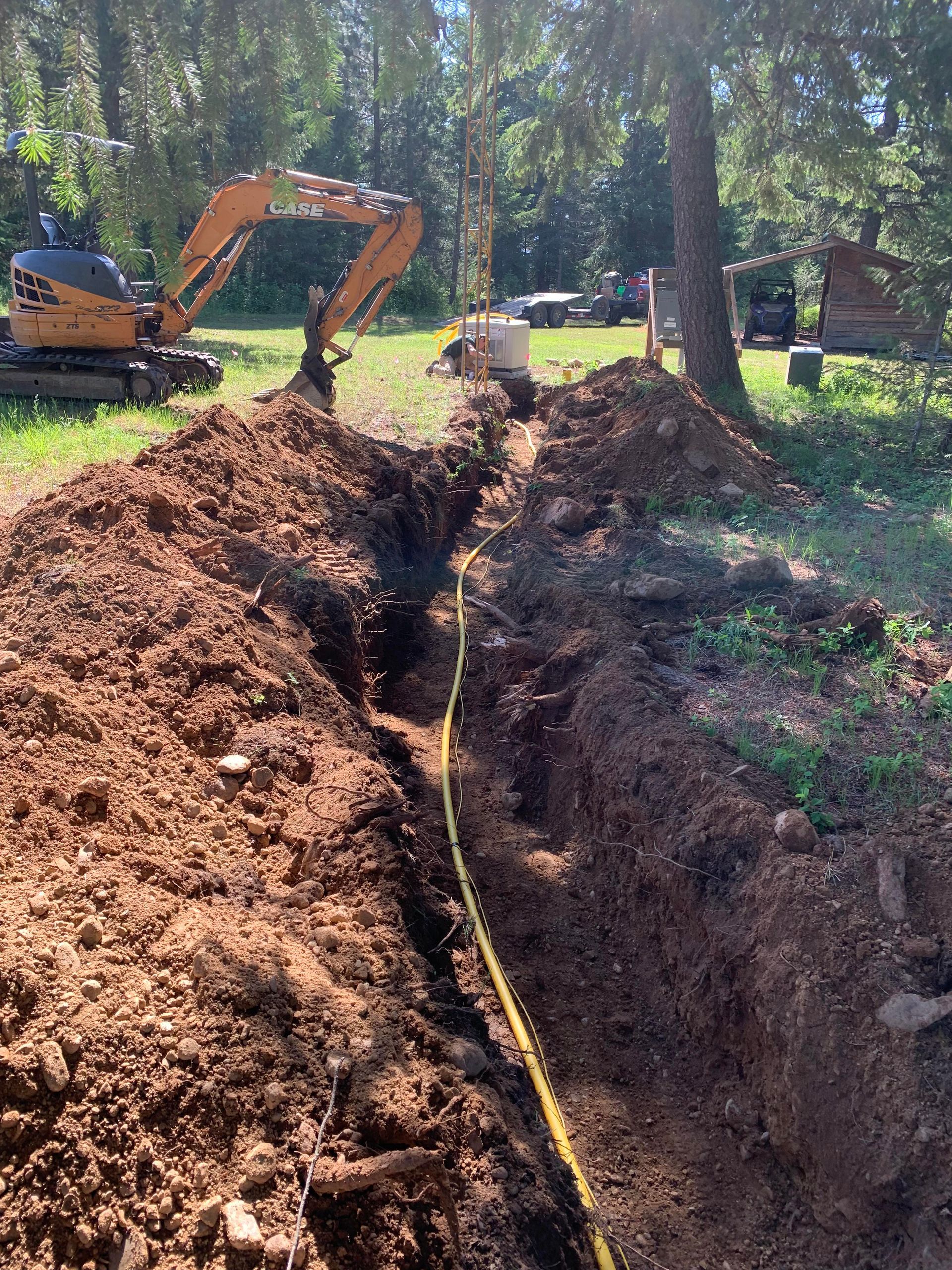 Trench dug in dirt with yellow cable; excavator in background, near trees.