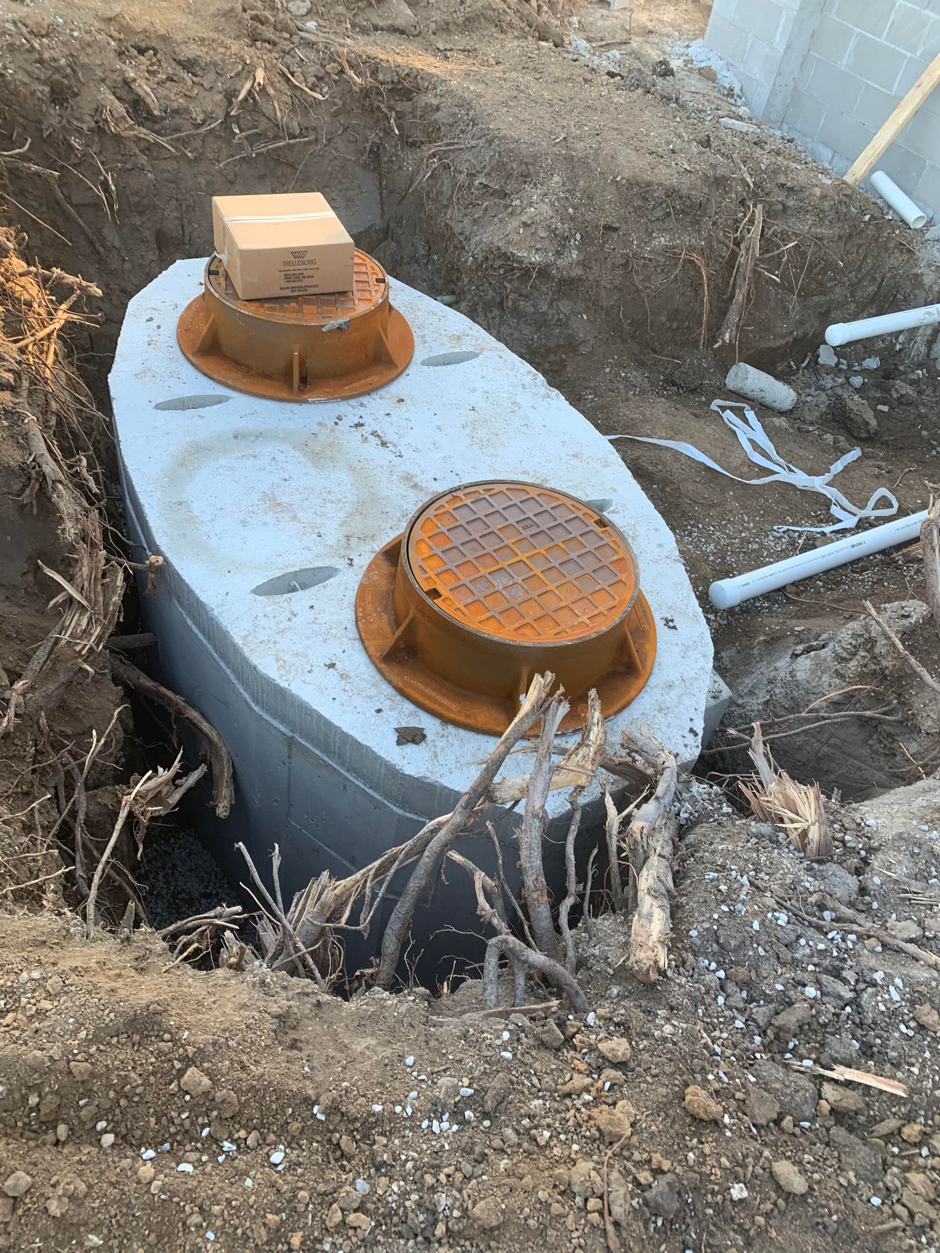 Concrete septic tank, with two rusty-orange lids, in an excavated pit; dirt, roots, and a cardboard box are visible.
