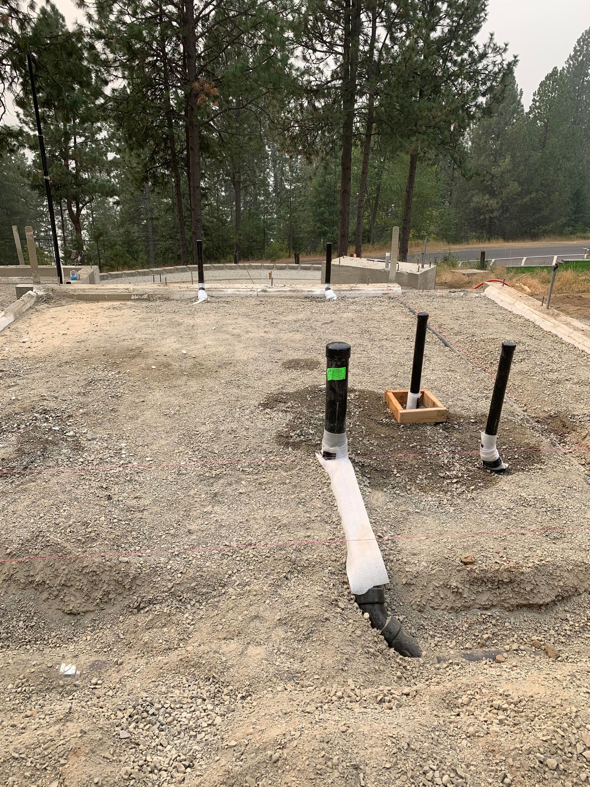 Construction site with gravel, plumbing pipes protruding, and forest background.