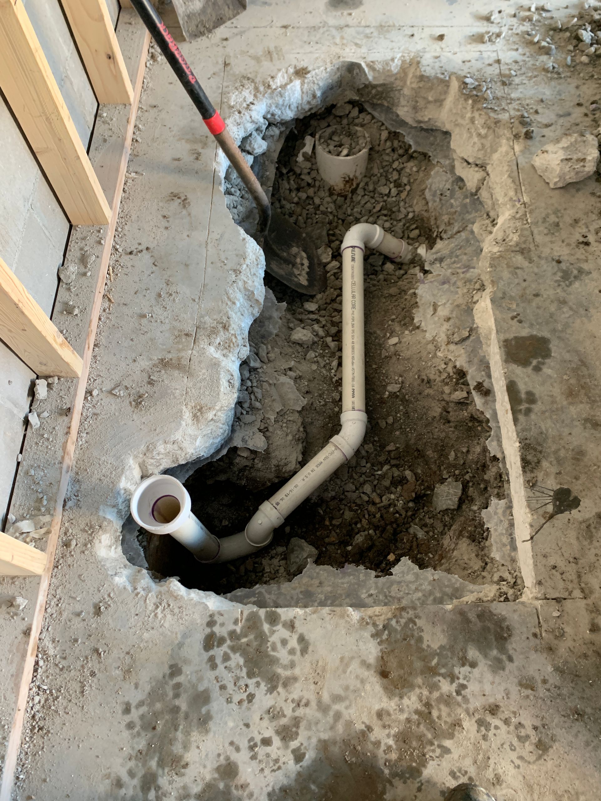 Piping exposed in a concrete floor opening, with a shovel; likely plumbing repair or installation in progress.