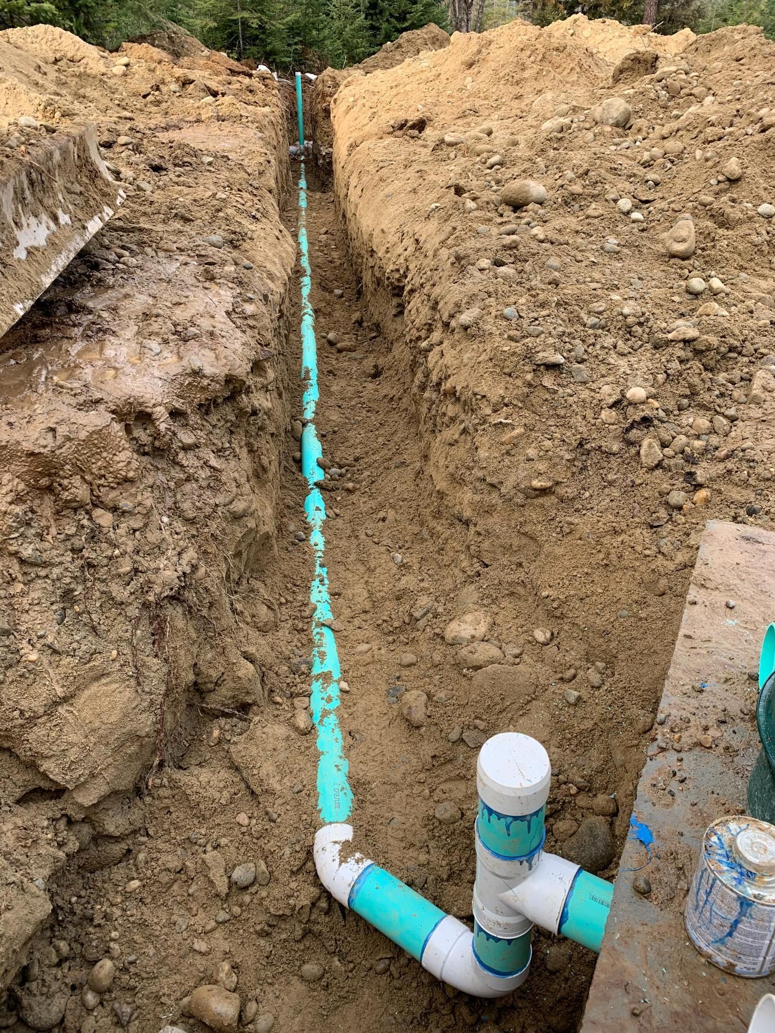 Trench with installed teal and white PVC pipes, likely for plumbing or drainage, in a dirt ground.