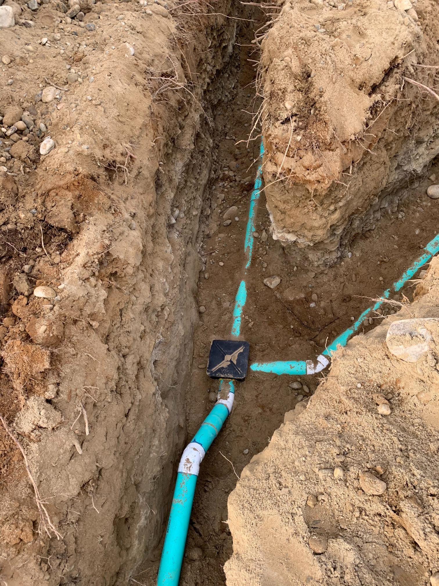 Trench in dirt with blue PVC pipes and connectors, some buried, and a black electrical box.