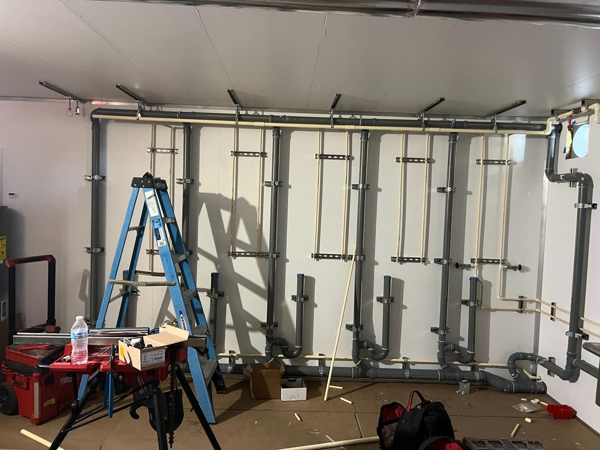 Construction of plumbing in a basement. PVC pipes and brackets are attached to the wall. A ladder and tools are present.