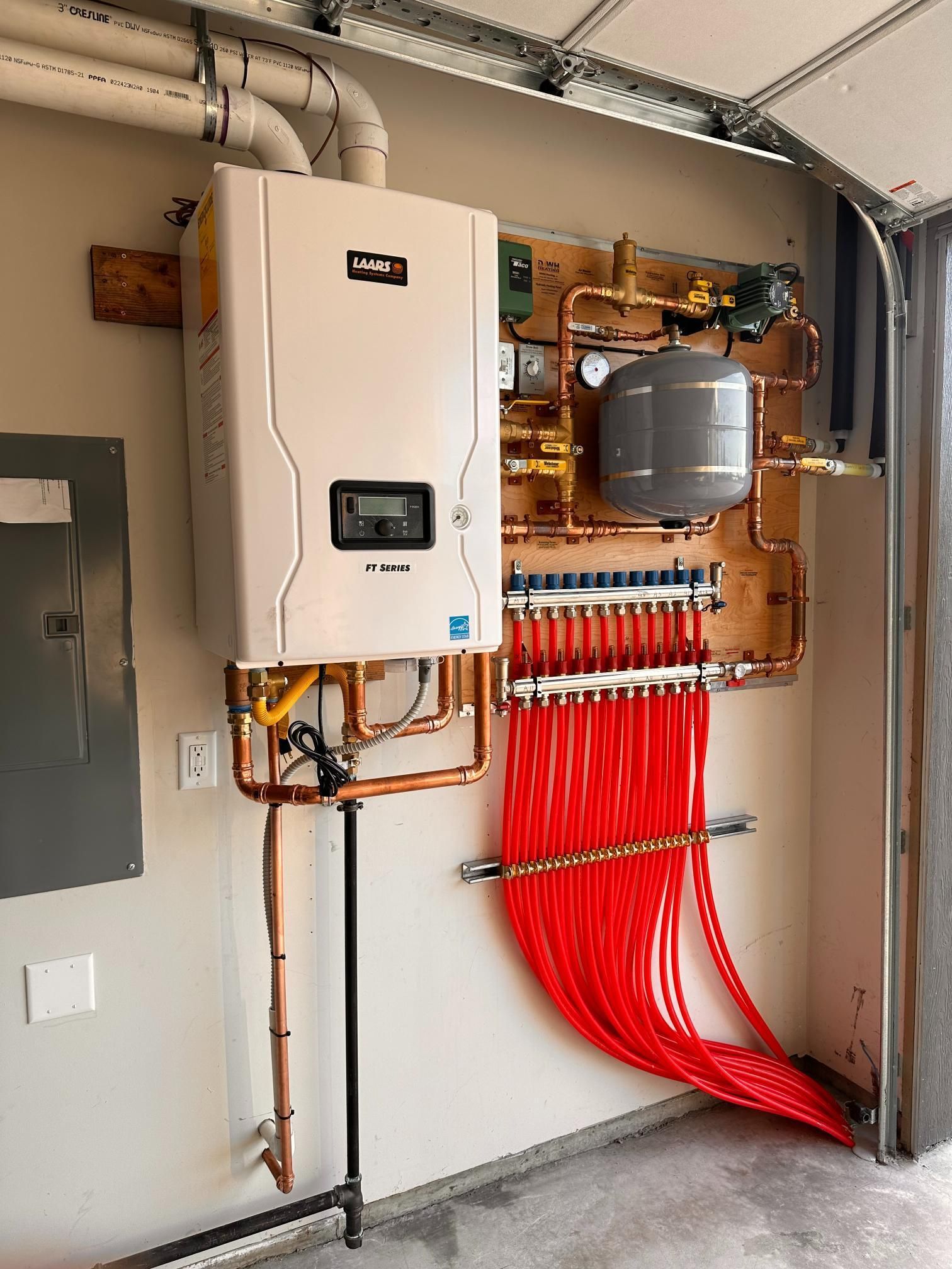 A wall-mounted, white tankless water heater with copper piping and red tubes, part of a radiant heating system.