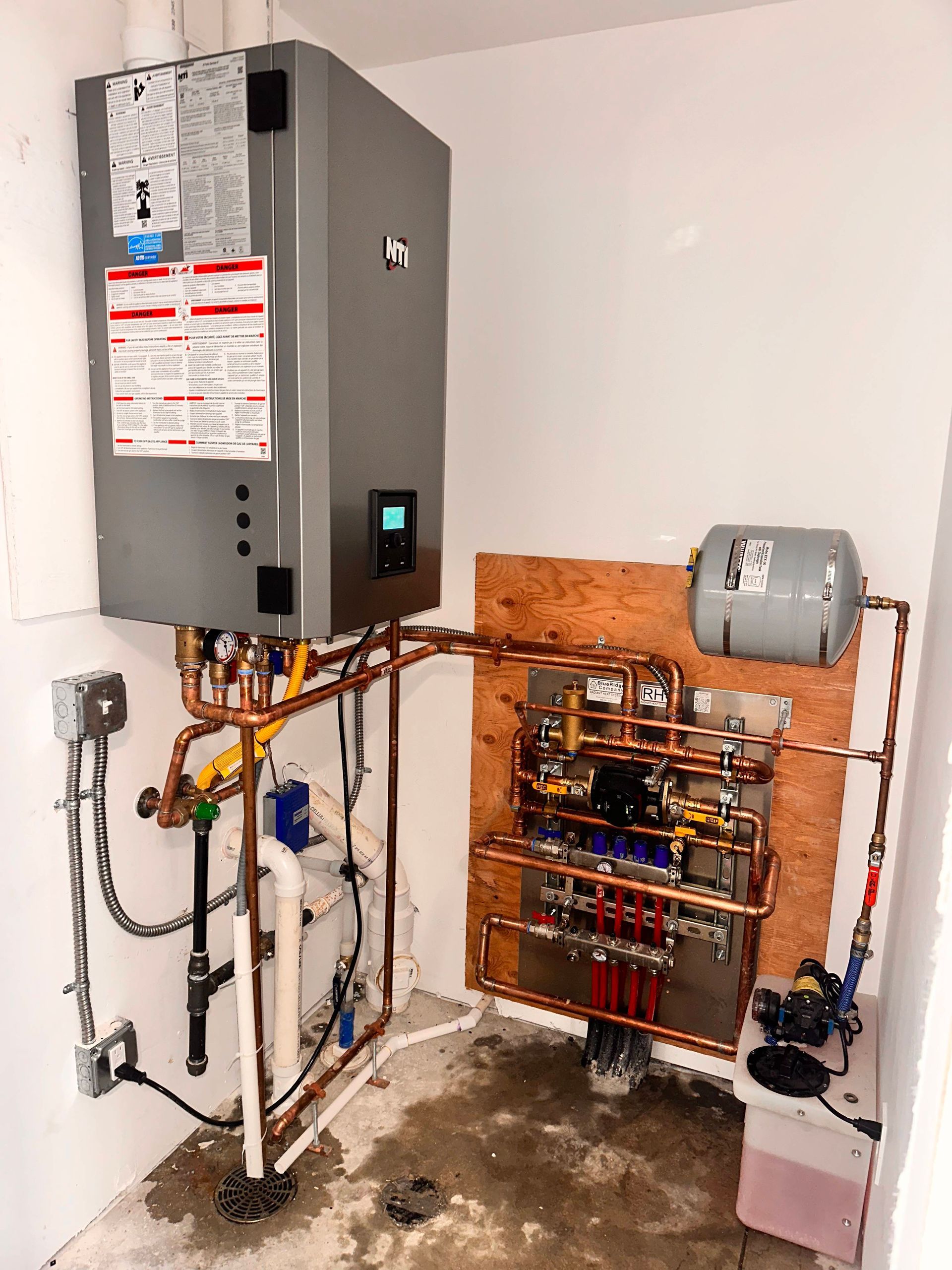 A grey wall-mounted boiler unit connected to copper piping, a control panel, and a small expansion tank in a utility room.