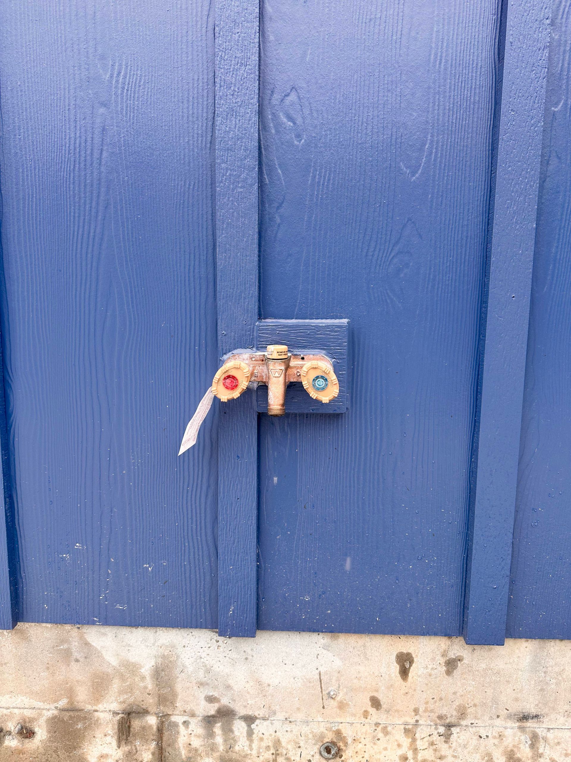 An outdoor dual-handle brass water spigot with red and blue accents, mounted on a blue wooden wall.