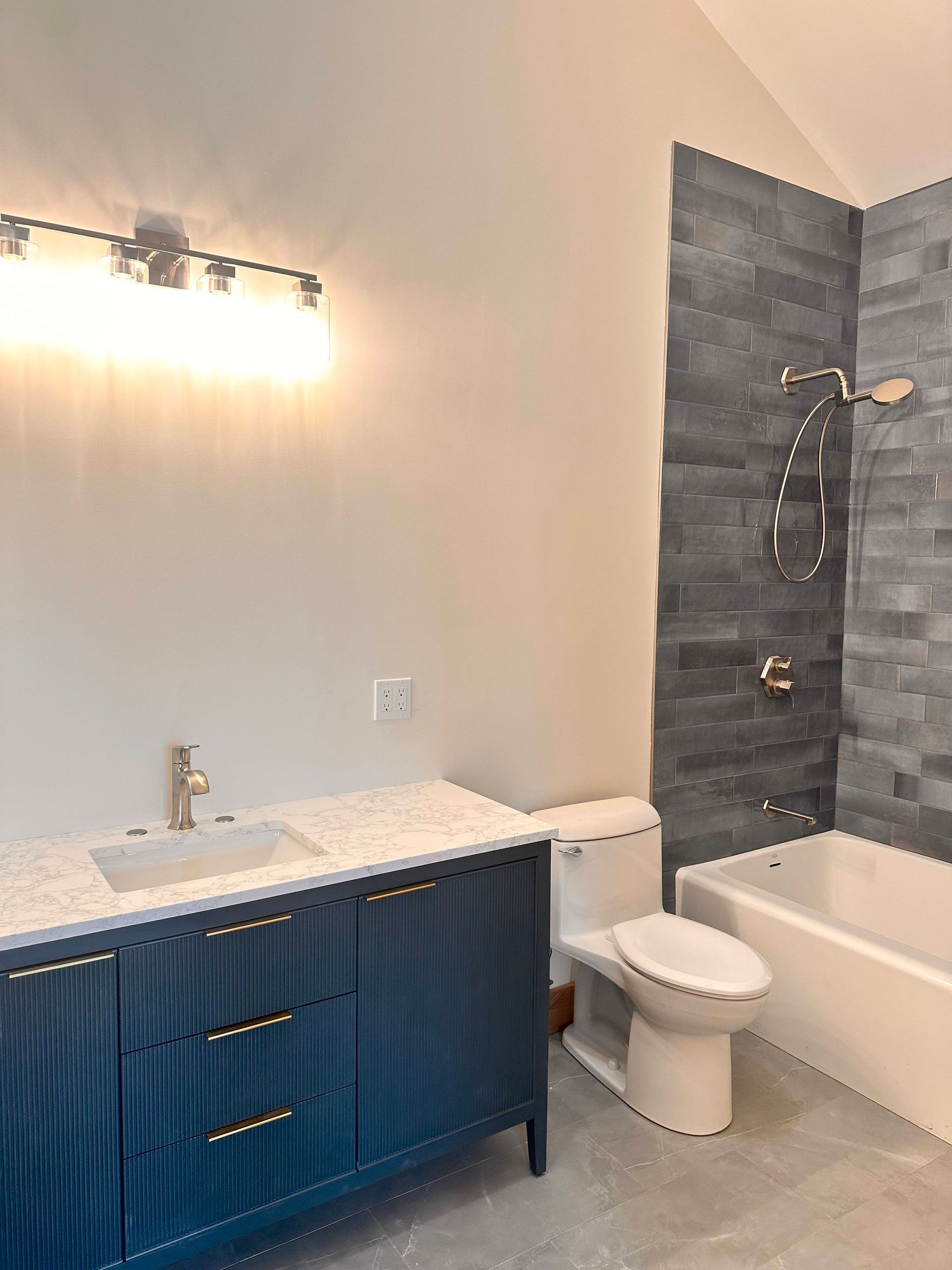 A modern bathroom featuring a dark blue vanity with gold hardware, a white sink, a toilet, and a grey tiled bathtub area.