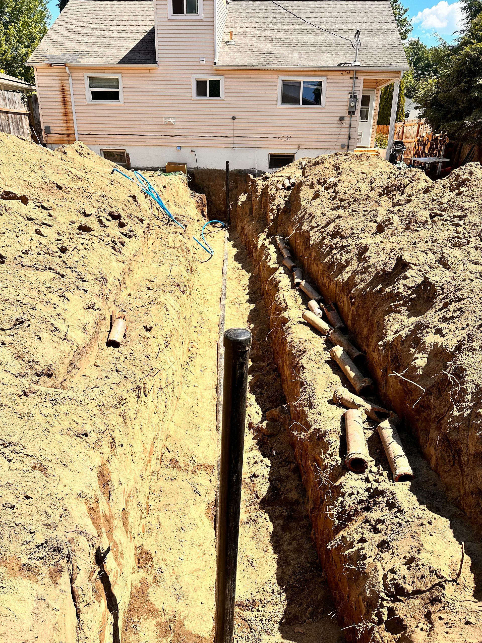 A deep trench dug in dirt with pipes visible; a house is in the background.