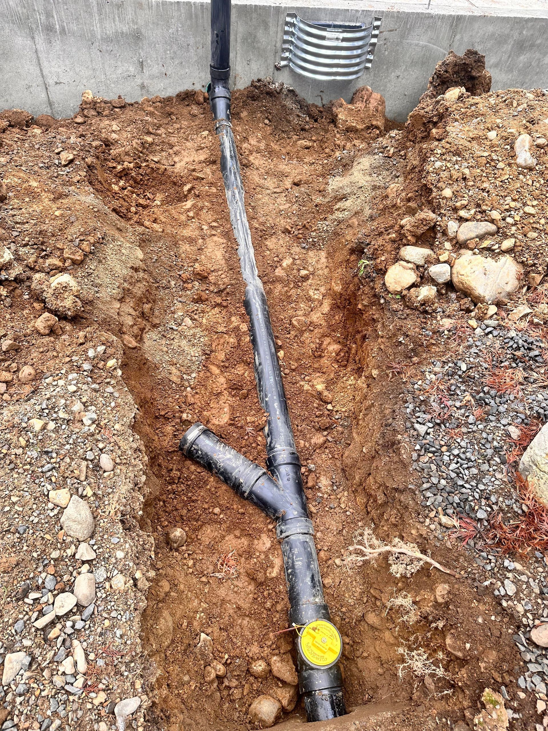 Black plumbing pipe in trench, connecting to a building's foundation, in a dirt setting.