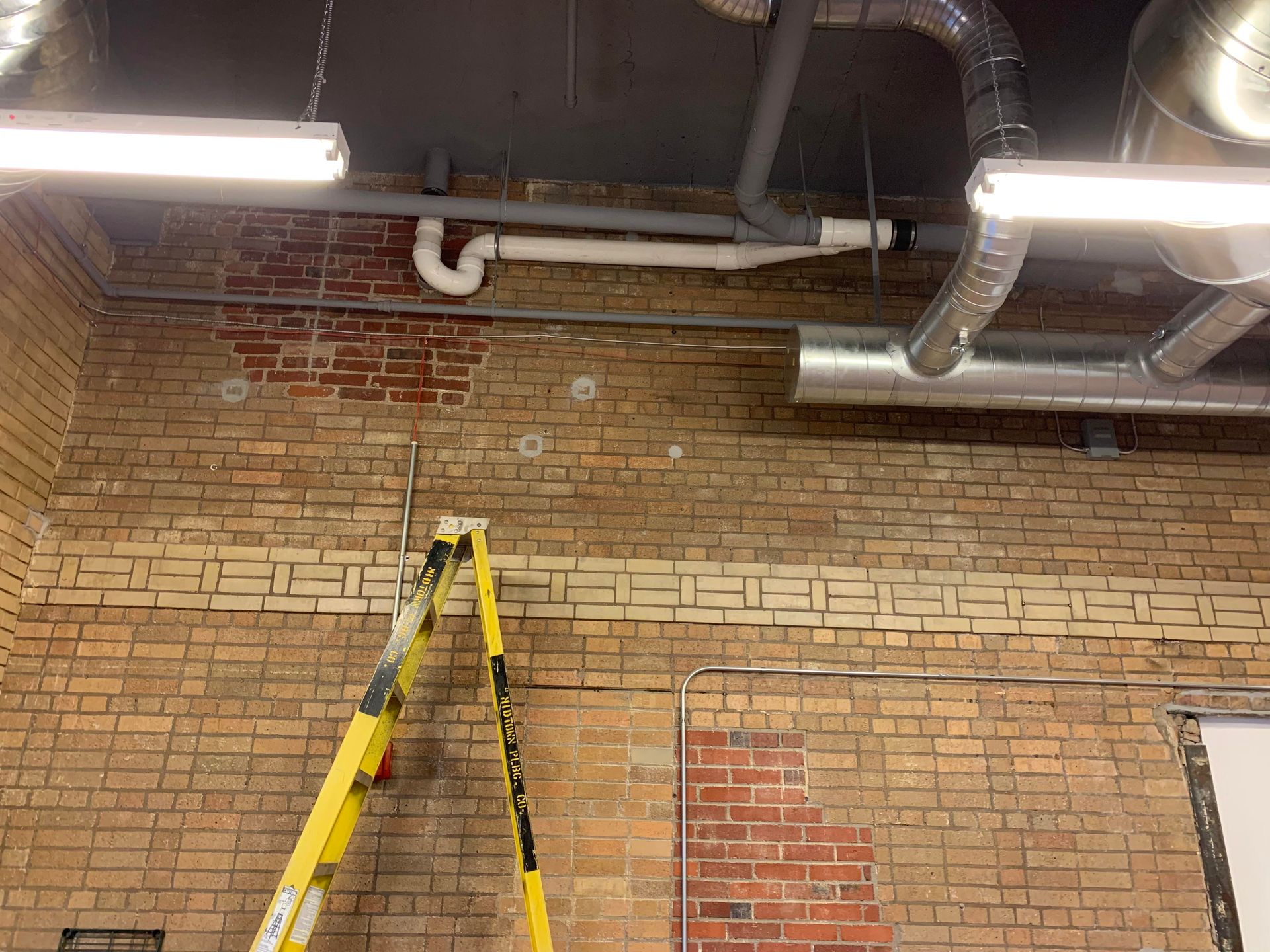 Brick wall with pipes and ventilation ducts against a dark ceiling, a ladder leans against the wall.