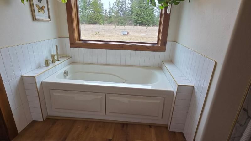 Bathroom with a white tiled tub, window overlooking field, wooden trim, wood floor.