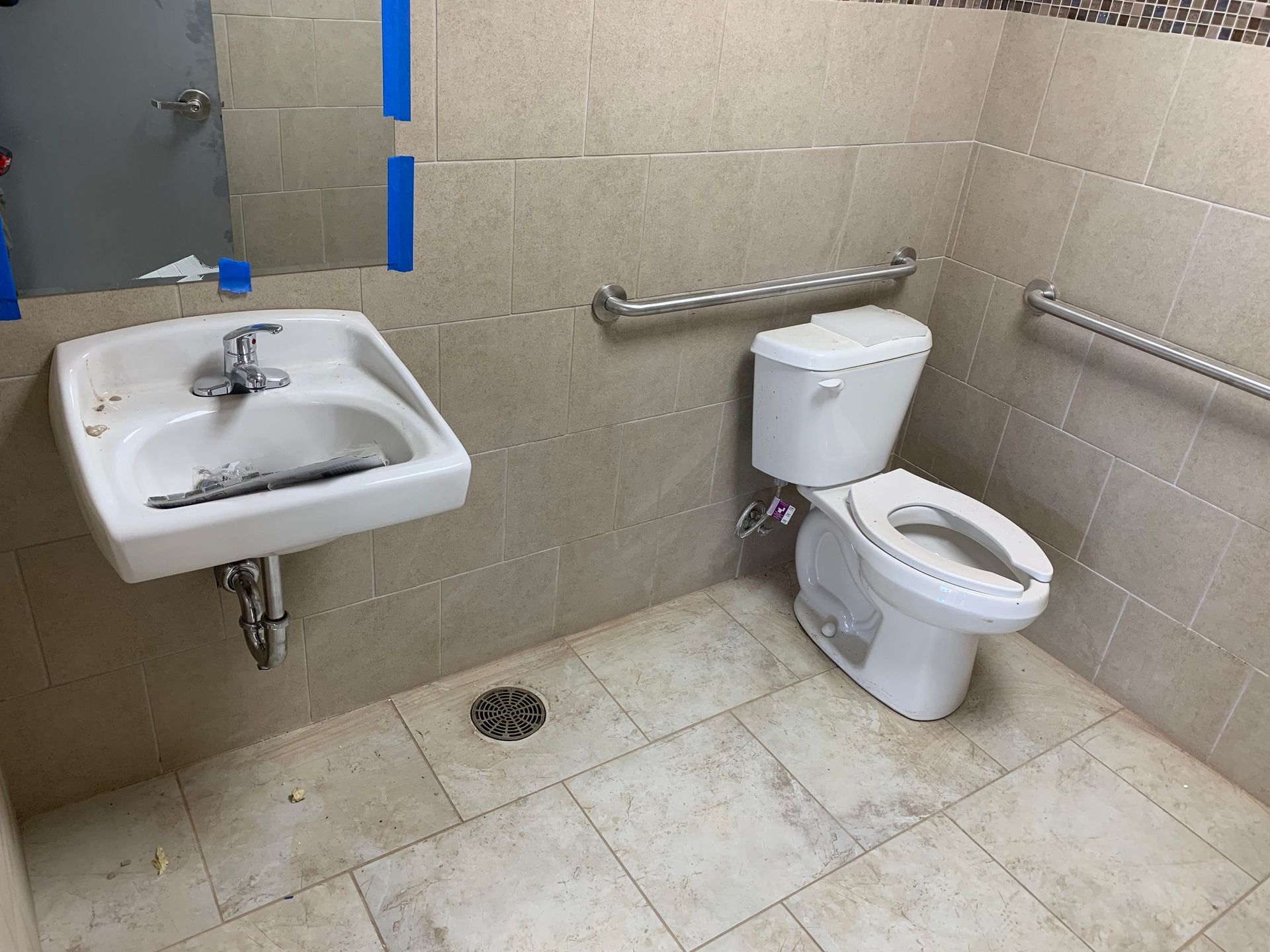 Public restroom with accessible toilet and sink. Beige tile, stainless steel grab bars, and a dirty sink.