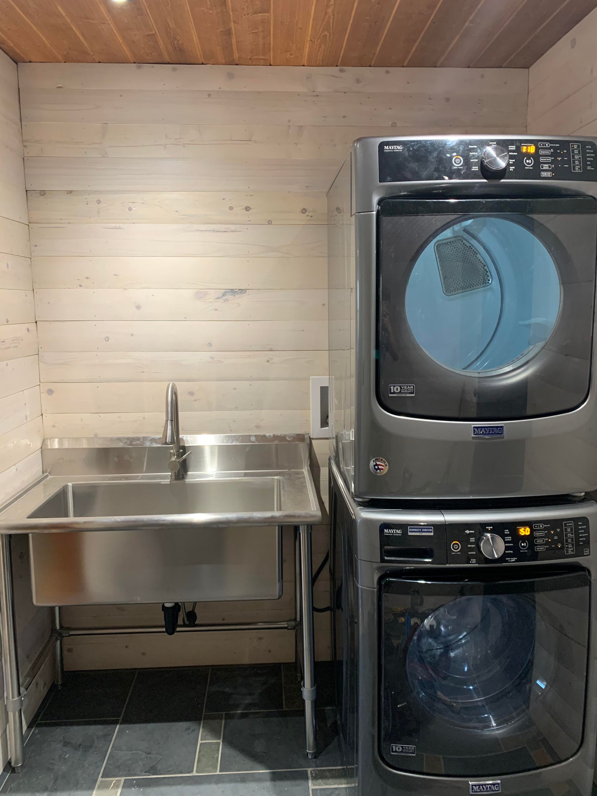 Laundry room with stacked washer and dryer, stainless steel sink, light wood paneling.