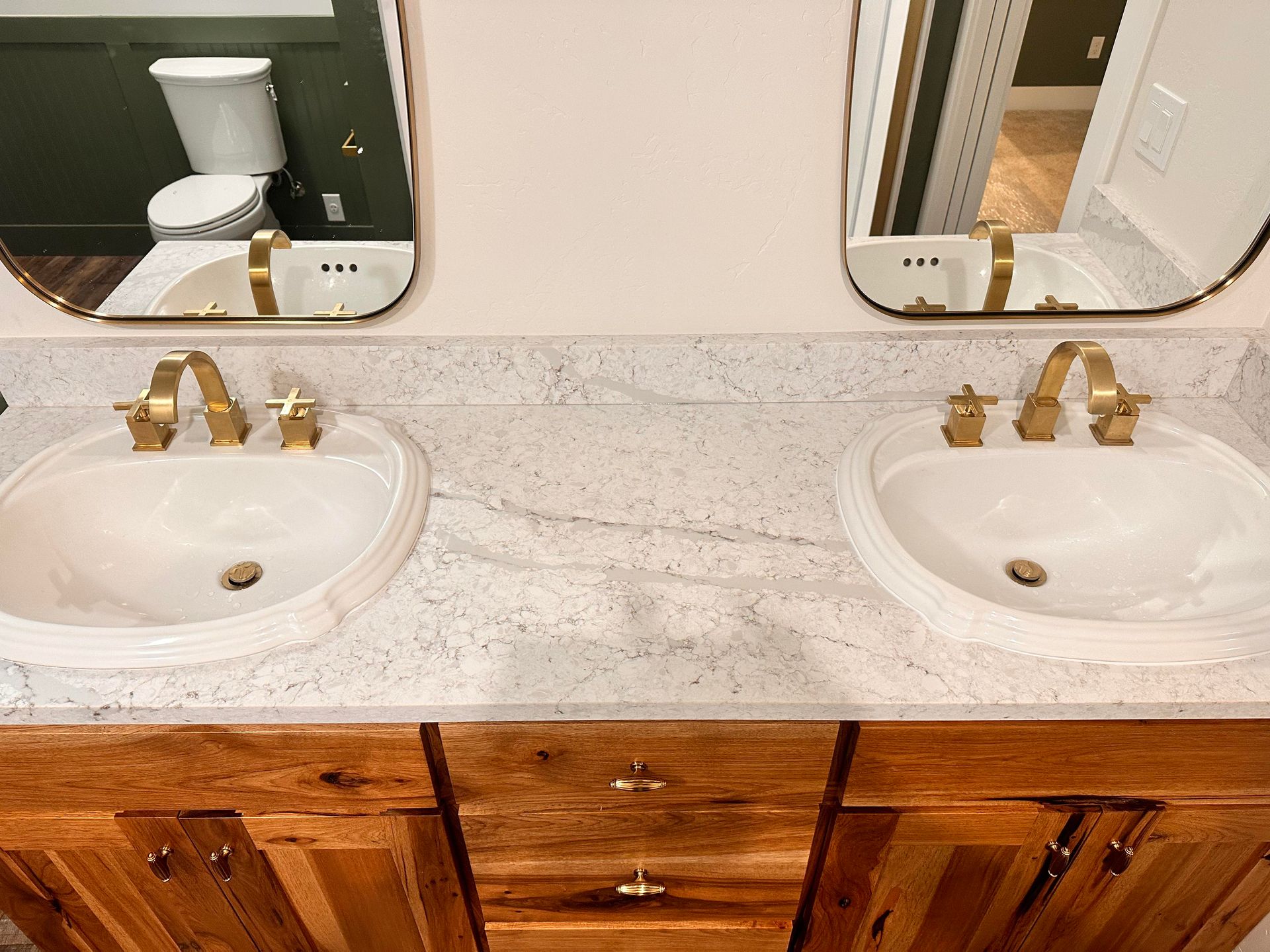 Bathroom with two sinks, gold faucets, and mirrors, on a white and gray countertop above a wooden vanity.