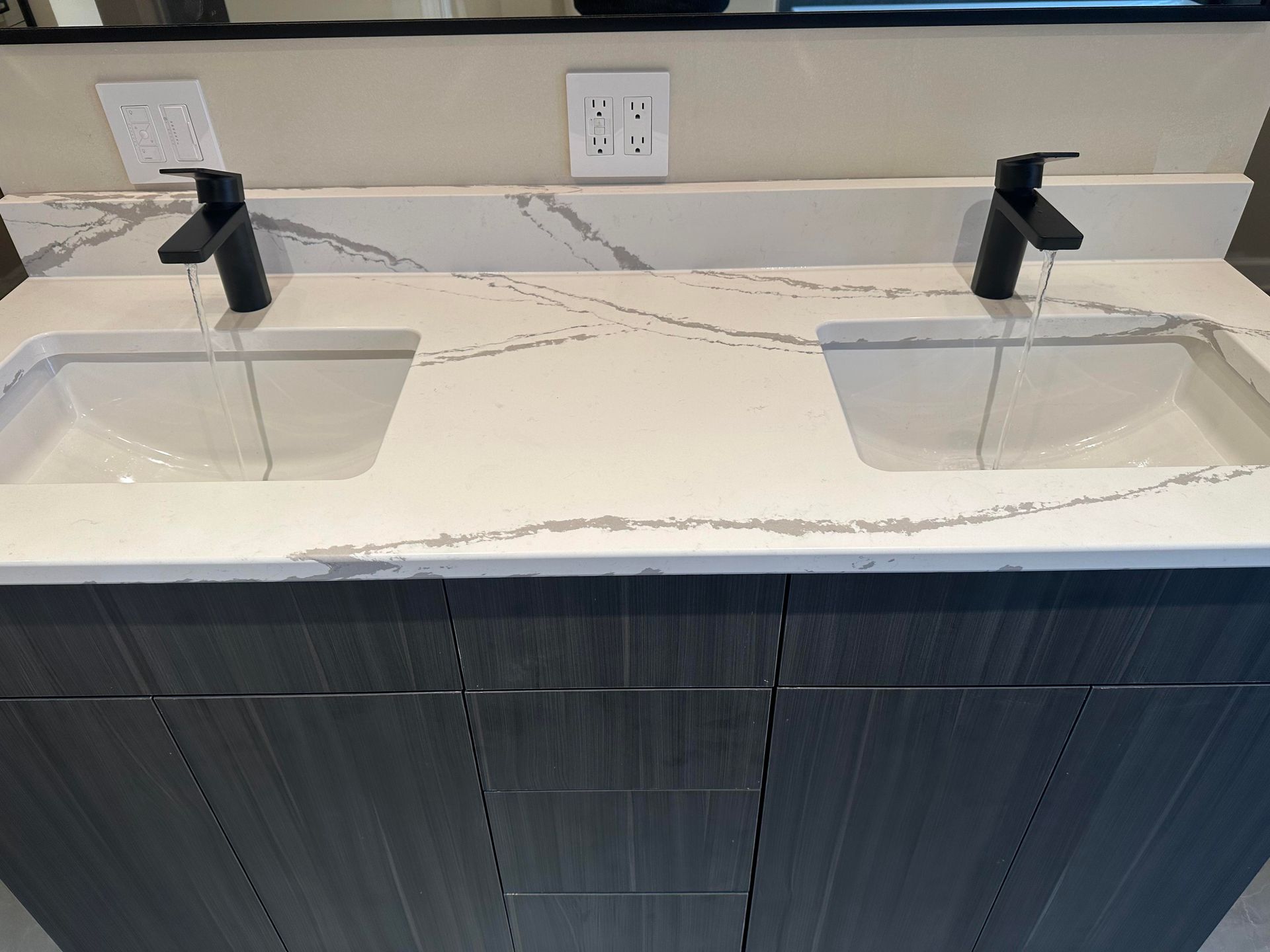 Double bathroom vanity with black faucets and white sinks; water running; gray countertop.