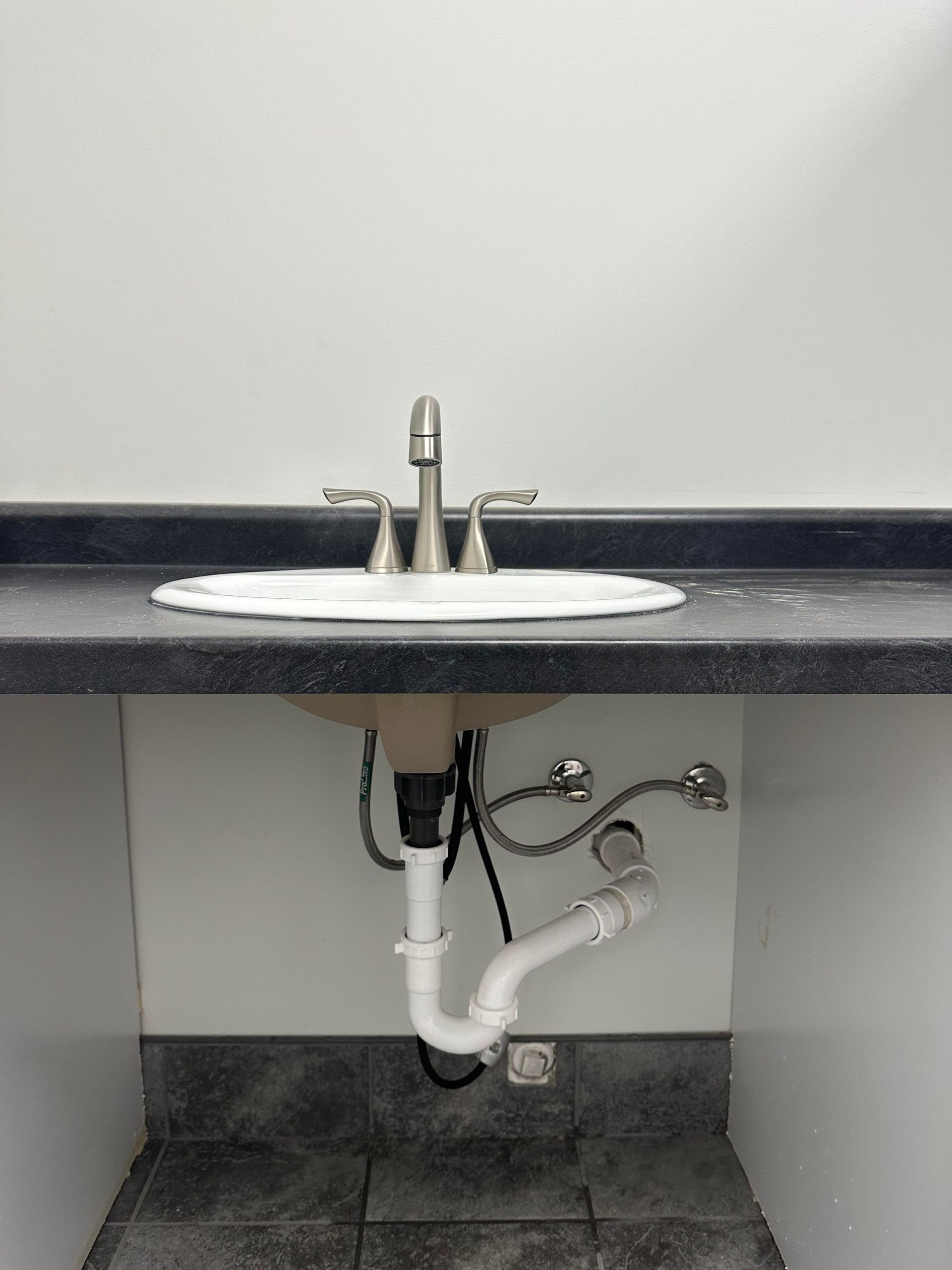 A bathroom sink with faucet and plumbing below, set in a white and black countertop, on tiled floor.