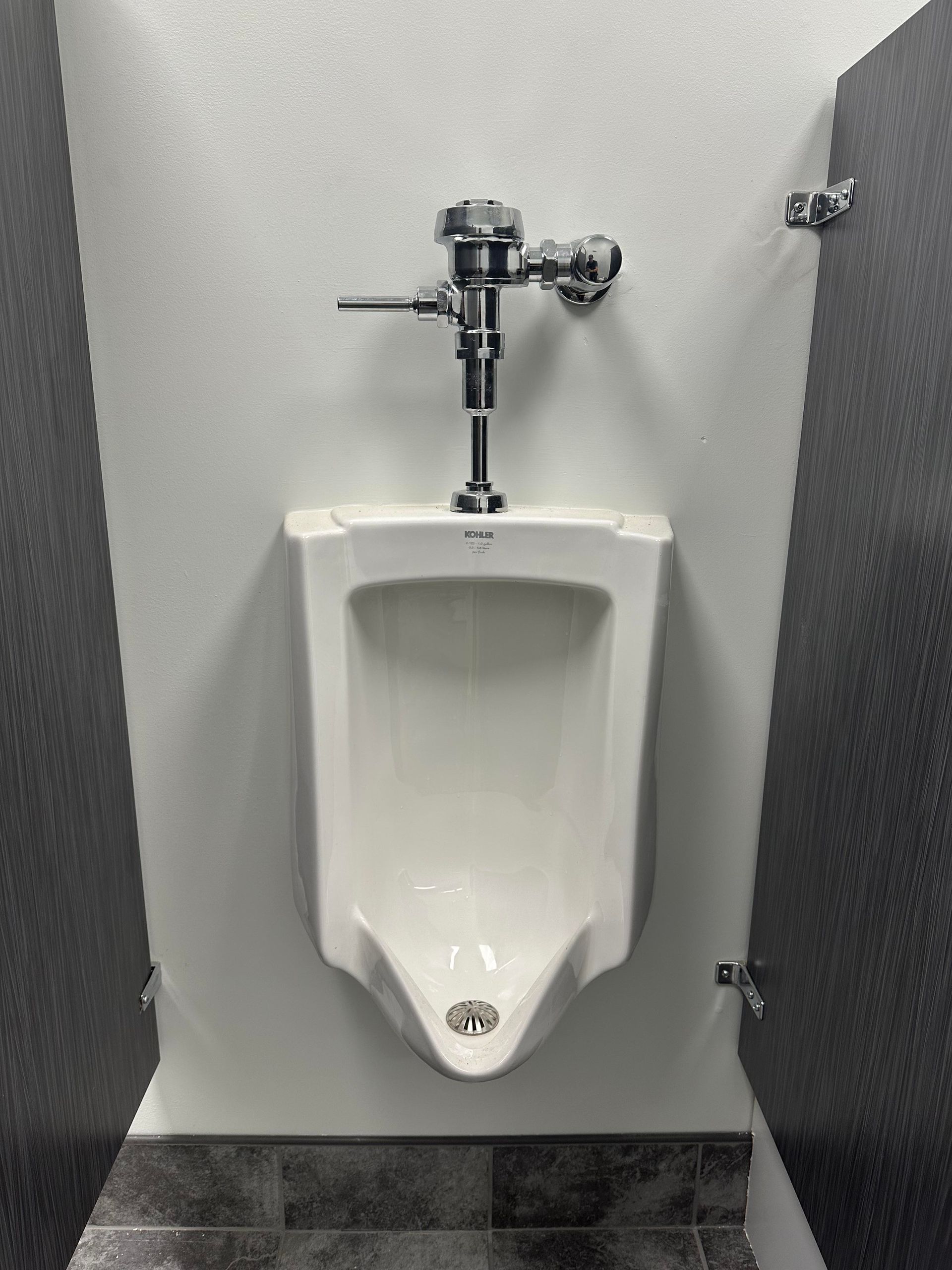 White urinal with chrome flush handle, in a bathroom stall with dark gray doors.