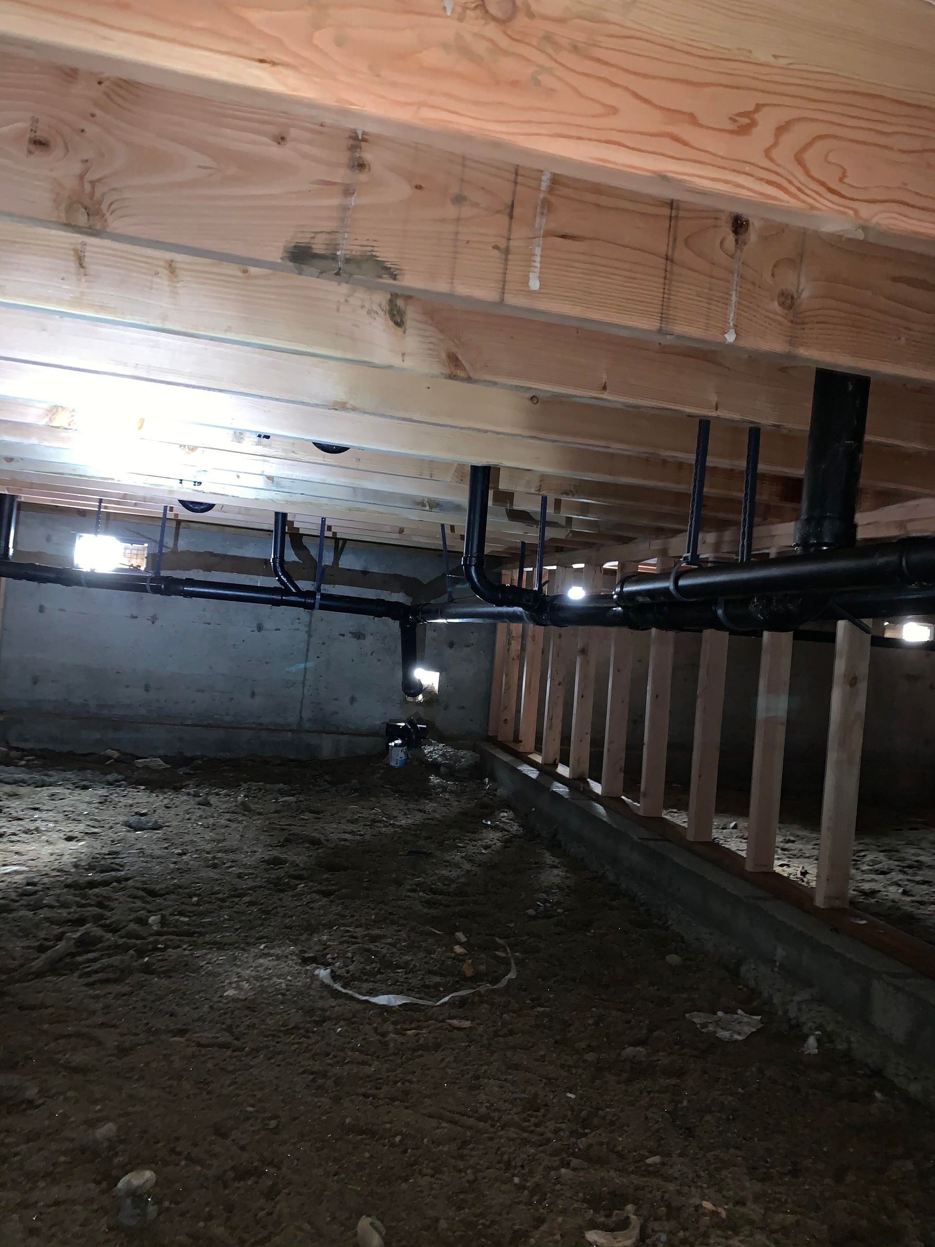 Underneath a house: exposed wooden beams, black pipes, dirt floor, concrete foundation, and wooden supports.