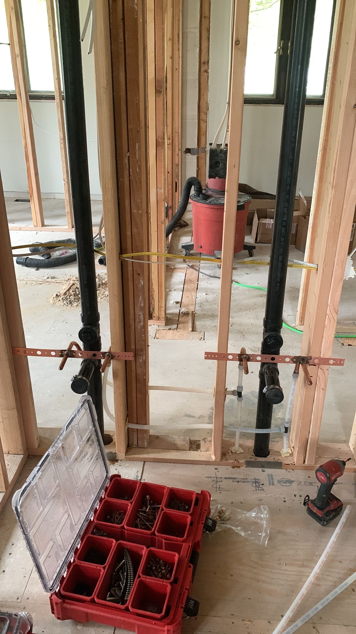 Construction site with plumbing pipes in wooden framing, tools on the floor, red organizer box.