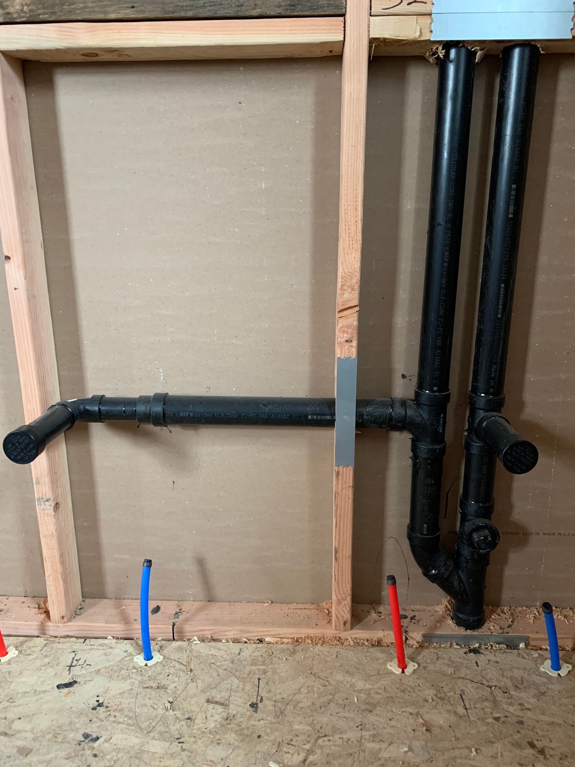 Plumbing for a bathroom: black pipes against a wooden frame, with blue and red markers on the floor.
