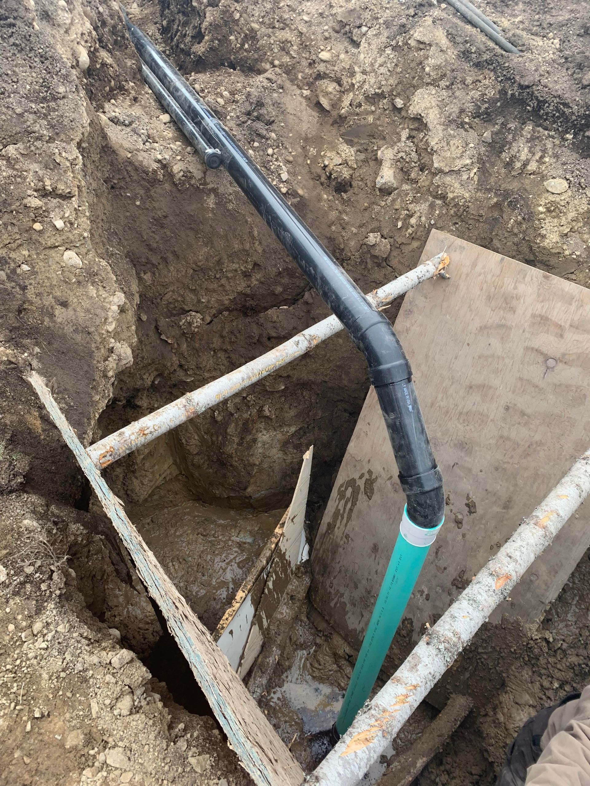 Black and teal pipes in a dirt trench. White support beams and plywood visible.