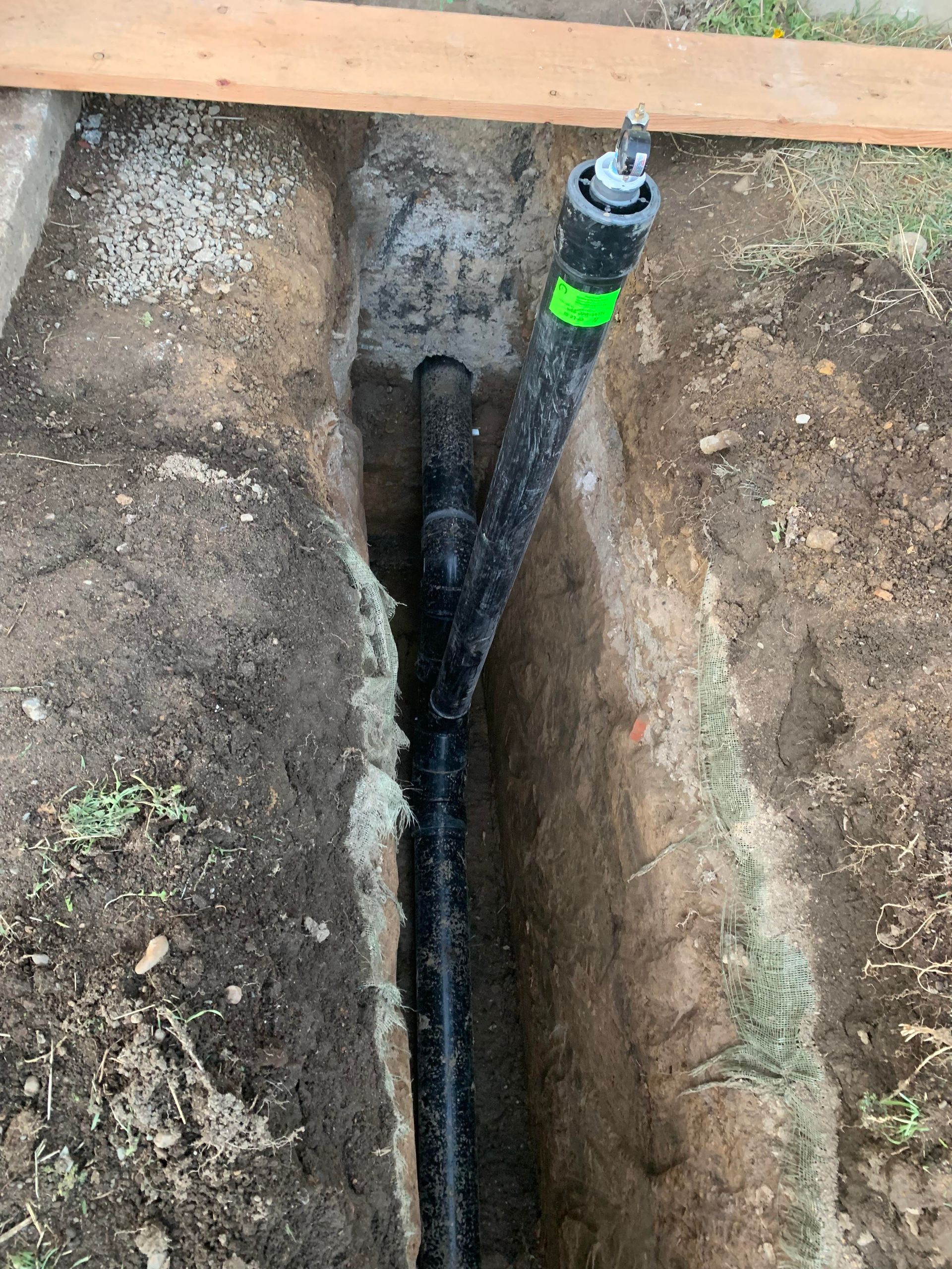 Black pipes in a trench, with a Y-connector and green tape. Dirt surrounds the plumbing.