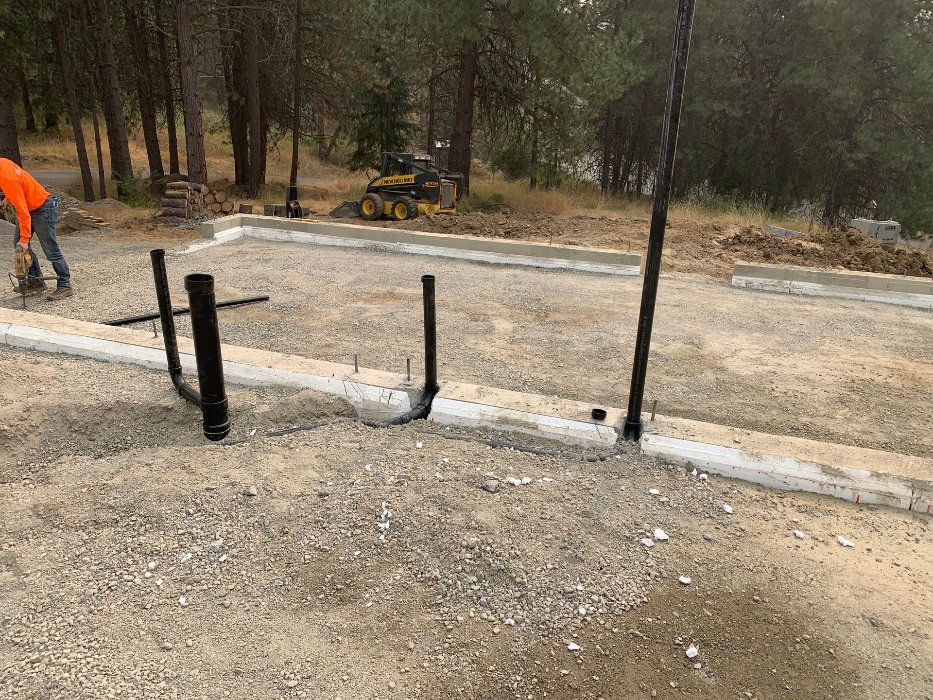 Black plumbing pipes rising from gravel. Man working in the background. Outdoors.