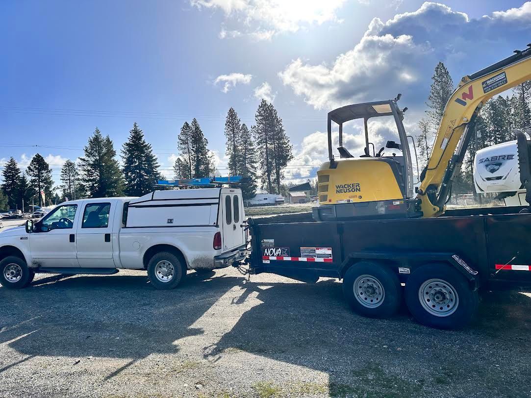 White truck towing a trailer with a yellow excavator on a gravel lot.