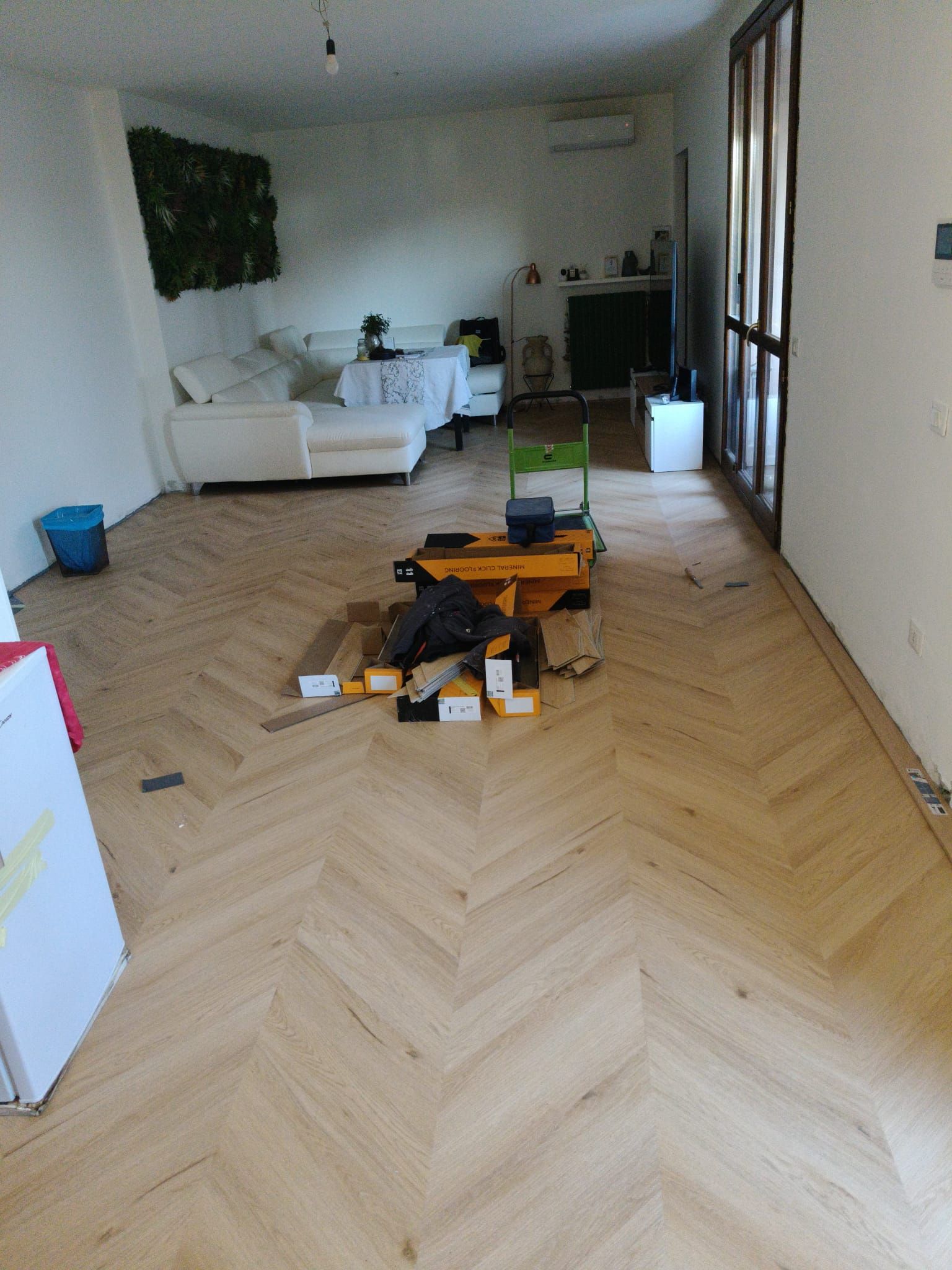 Soggiorno con pavimento in legno a spina di pesce e attrezzi sparsi, visto dall'ingresso.