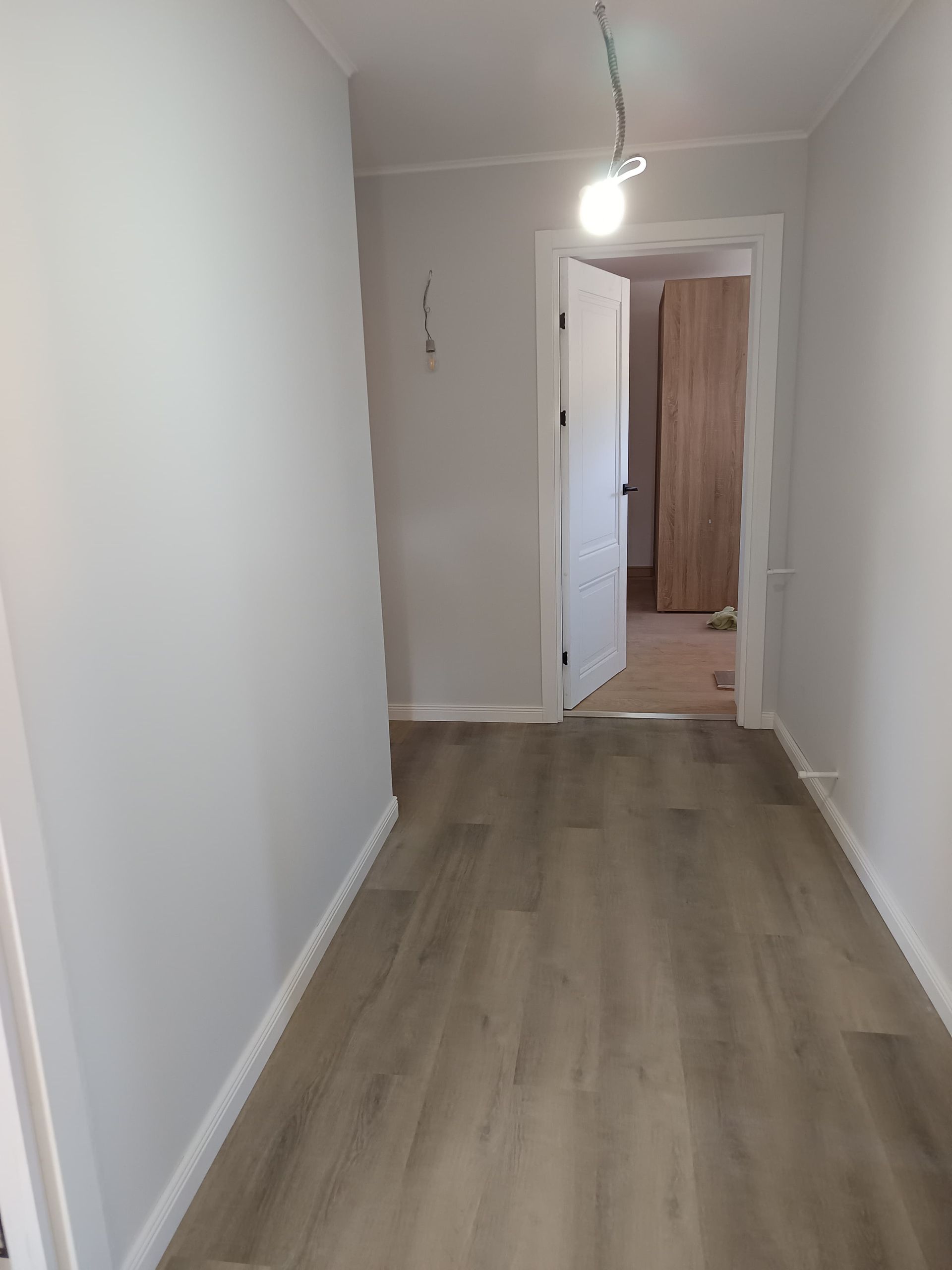 Stretto corridoio con pareti bianche, pavimento in legno chiaro e porta aperta in fondo.