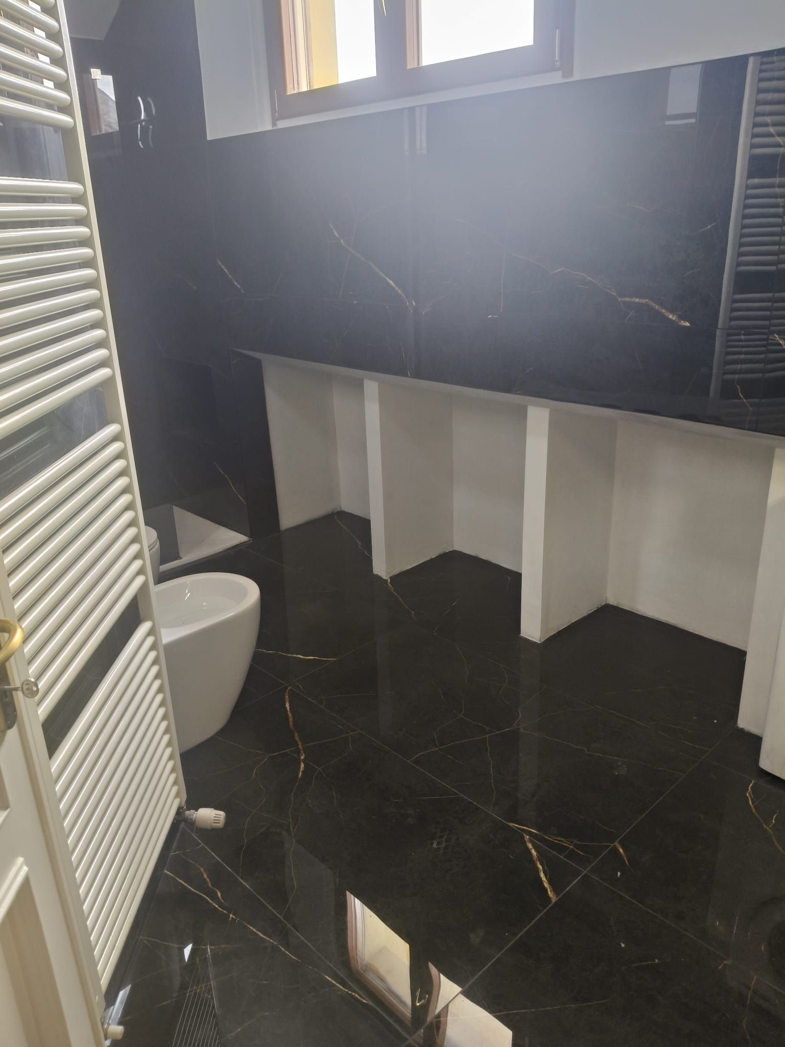 Piccolo bagno con pavimento in piastrelle nere, mobiletto bianco, WC e finestra sopra il lavabo.