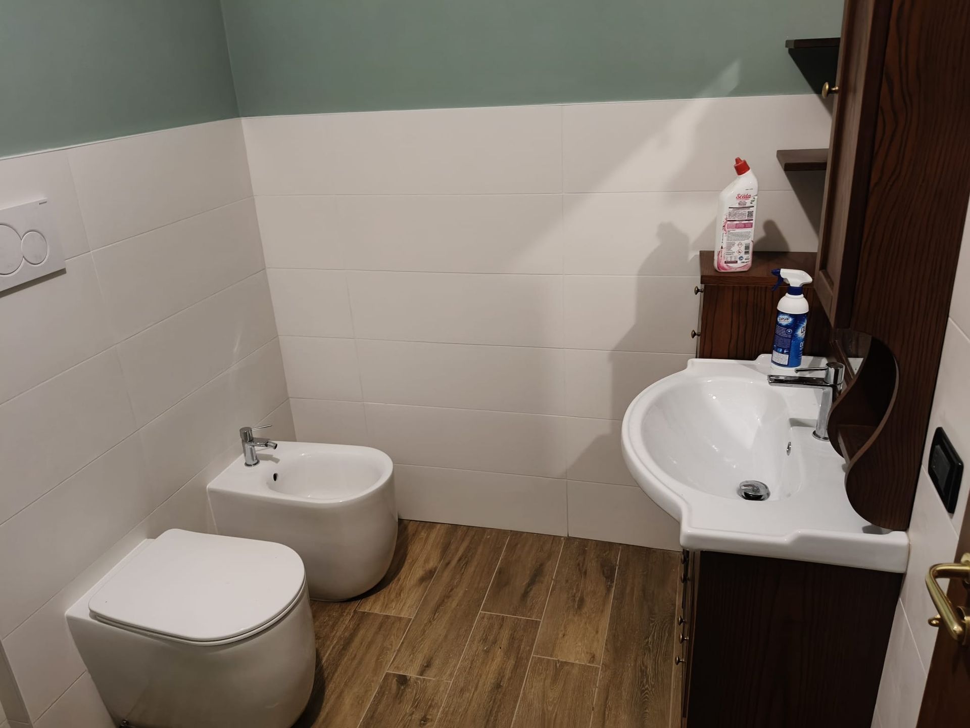 Piccolo bagno con WC, bidet, lavabo bianco e pavimento effetto legno.