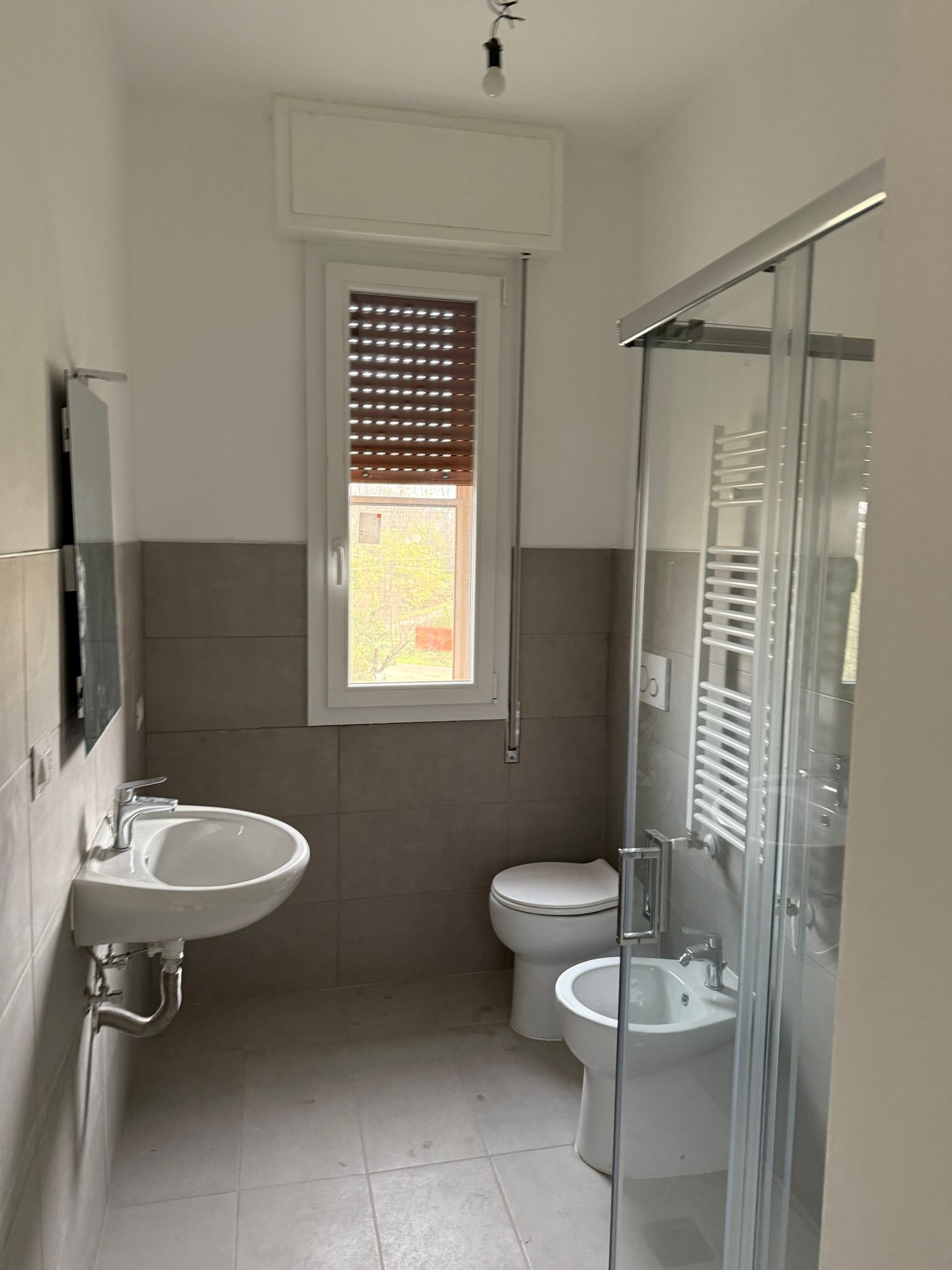 Piccolo bagno con lavabo, WC, bidet e box doccia in vetro; piastrelle grigie e una finestra con tenda a rullo.