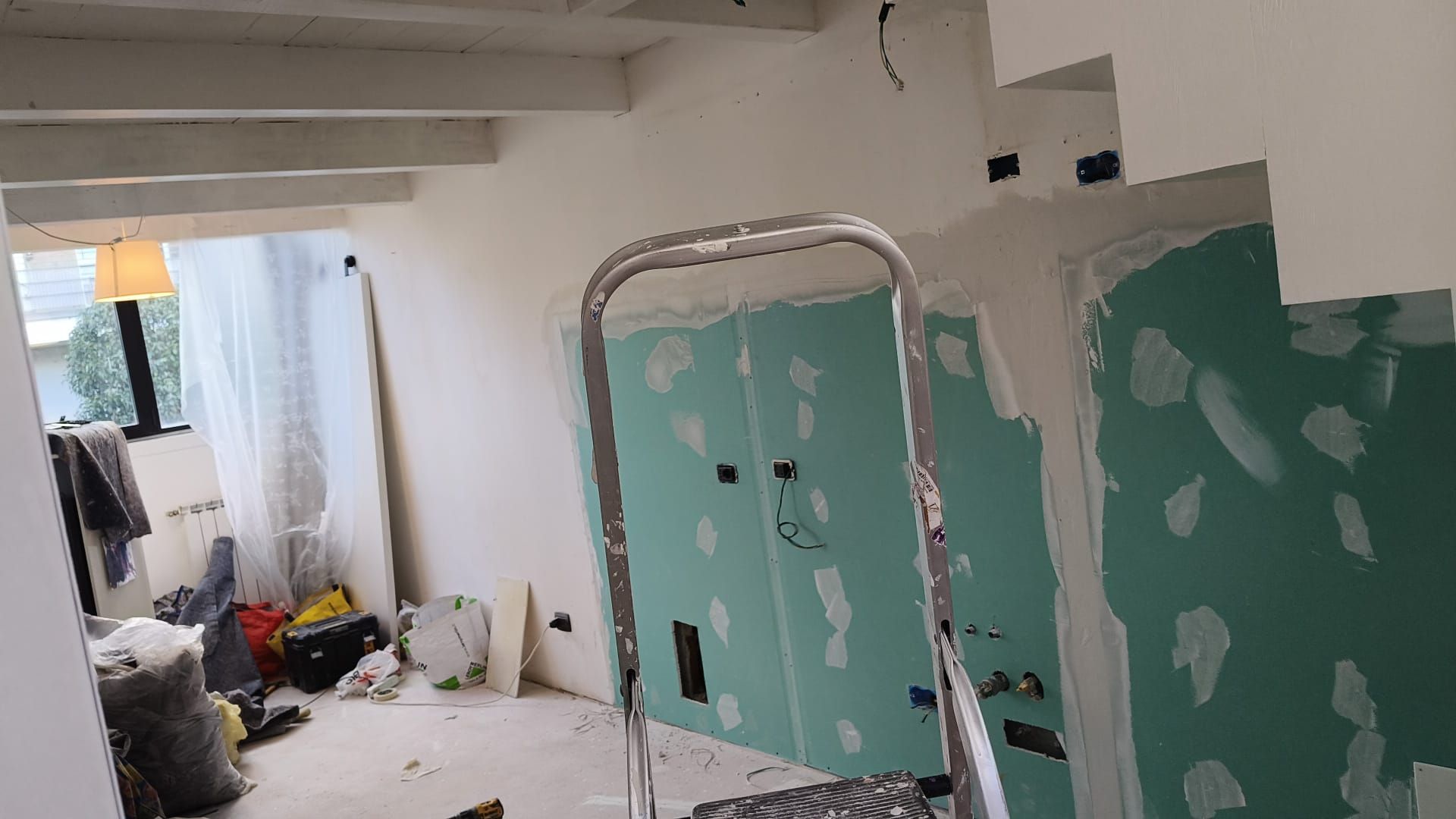 Stanza parzialmente ristrutturata con chiazze di verde sulle pareti, una scaletta e materiali edili sul pavimento.