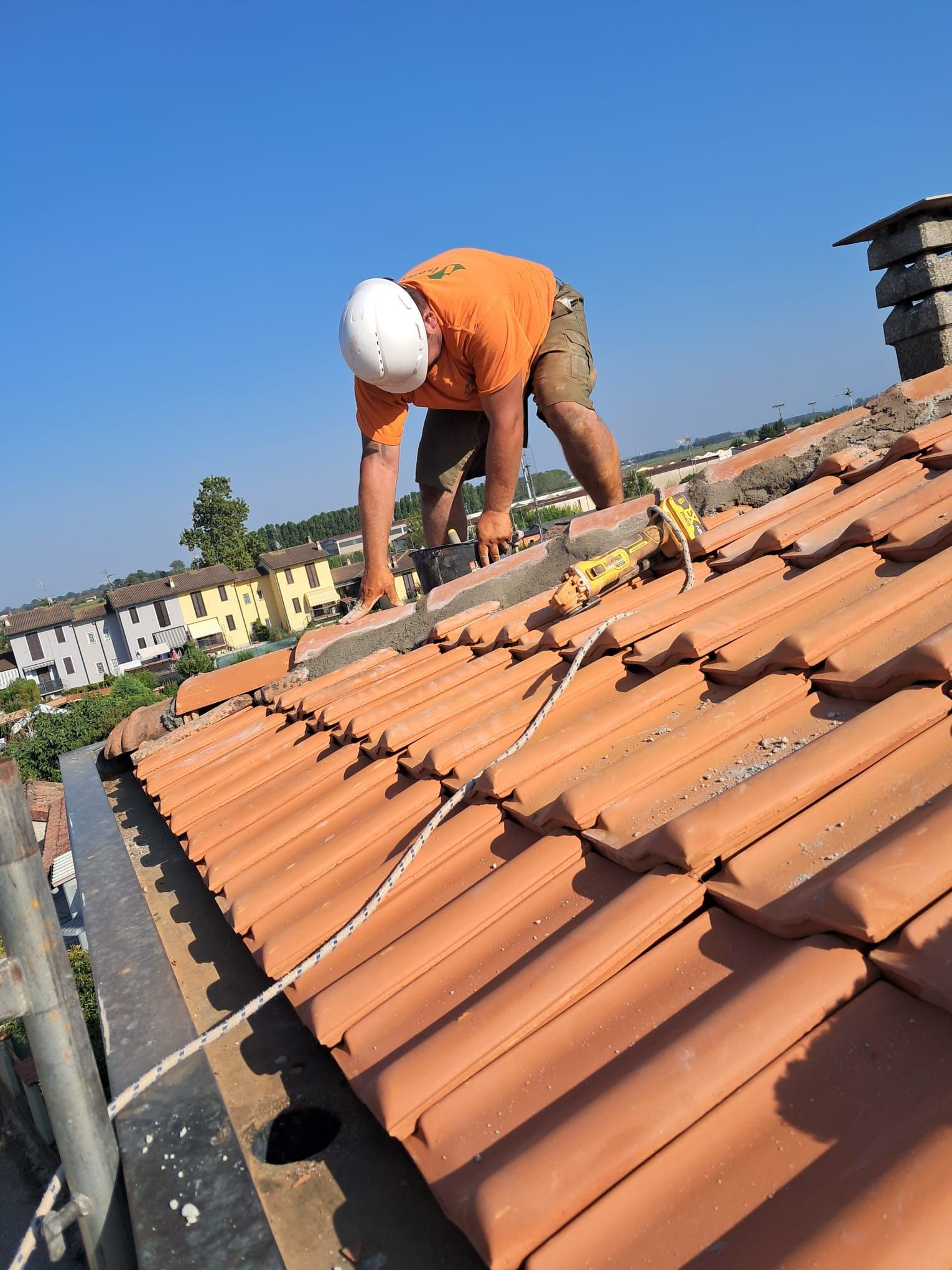 Un operaio con camicia arancione e casco bianco ripara delle tegole in terracotta su un tetto inclinato.