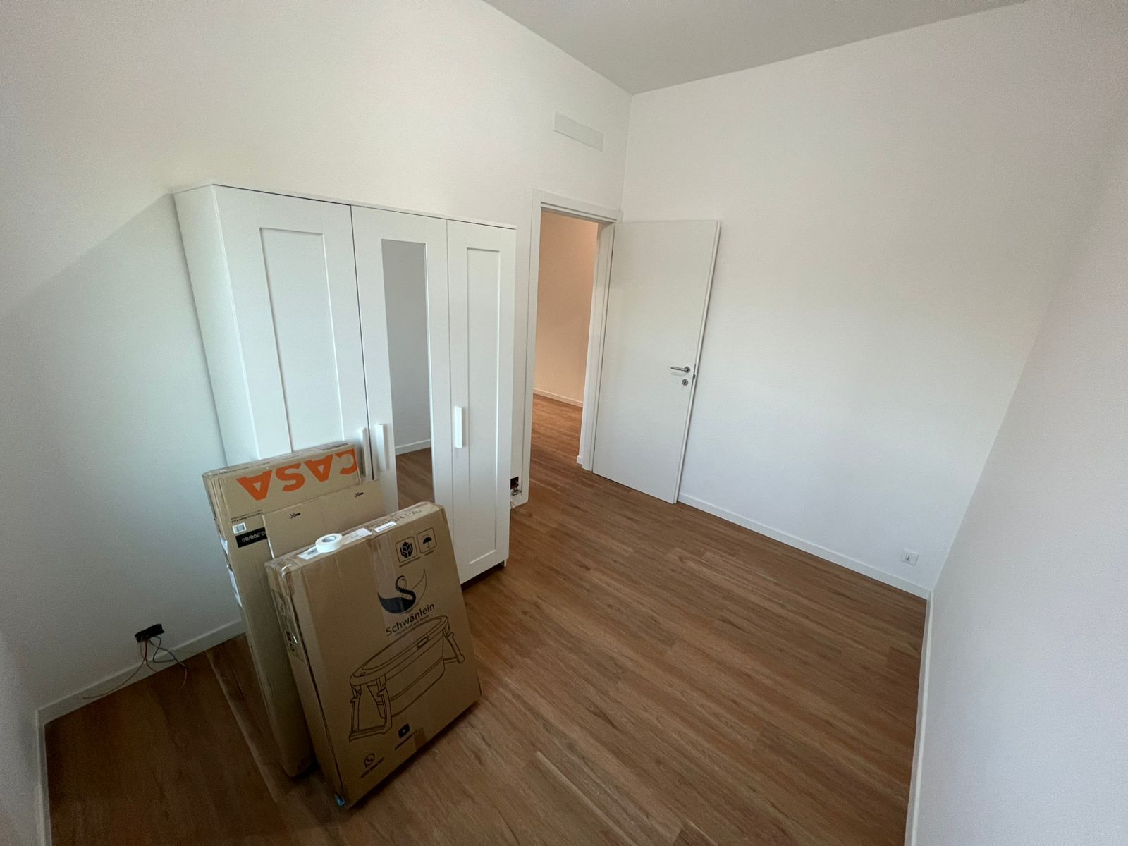 Stanza vuota con pareti bianche, pavimento in legno e armadio bianco; delle scatole sono appoggiate sulla porta.