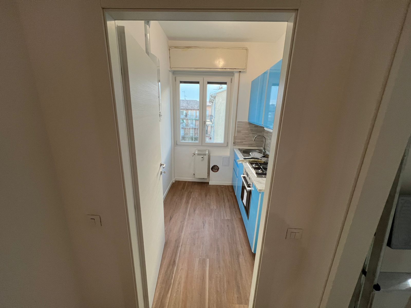 Stretto corridoio della cucina con pavimento in legno, mobili bianchi, elettrodomestici blu e una finestra in fondo.