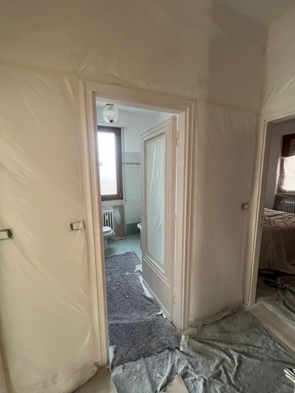 Corridoio coperto di plastica che conduce a una stanza luminosa durante i lavori di ristrutturazione, con porta e rivestimenti protettivi sul pavimento.